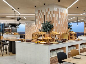 A modern cafe with a central island displaying pastries and bread, surrounded by seating. - Novotel Valencia Lavant (Valencia)
