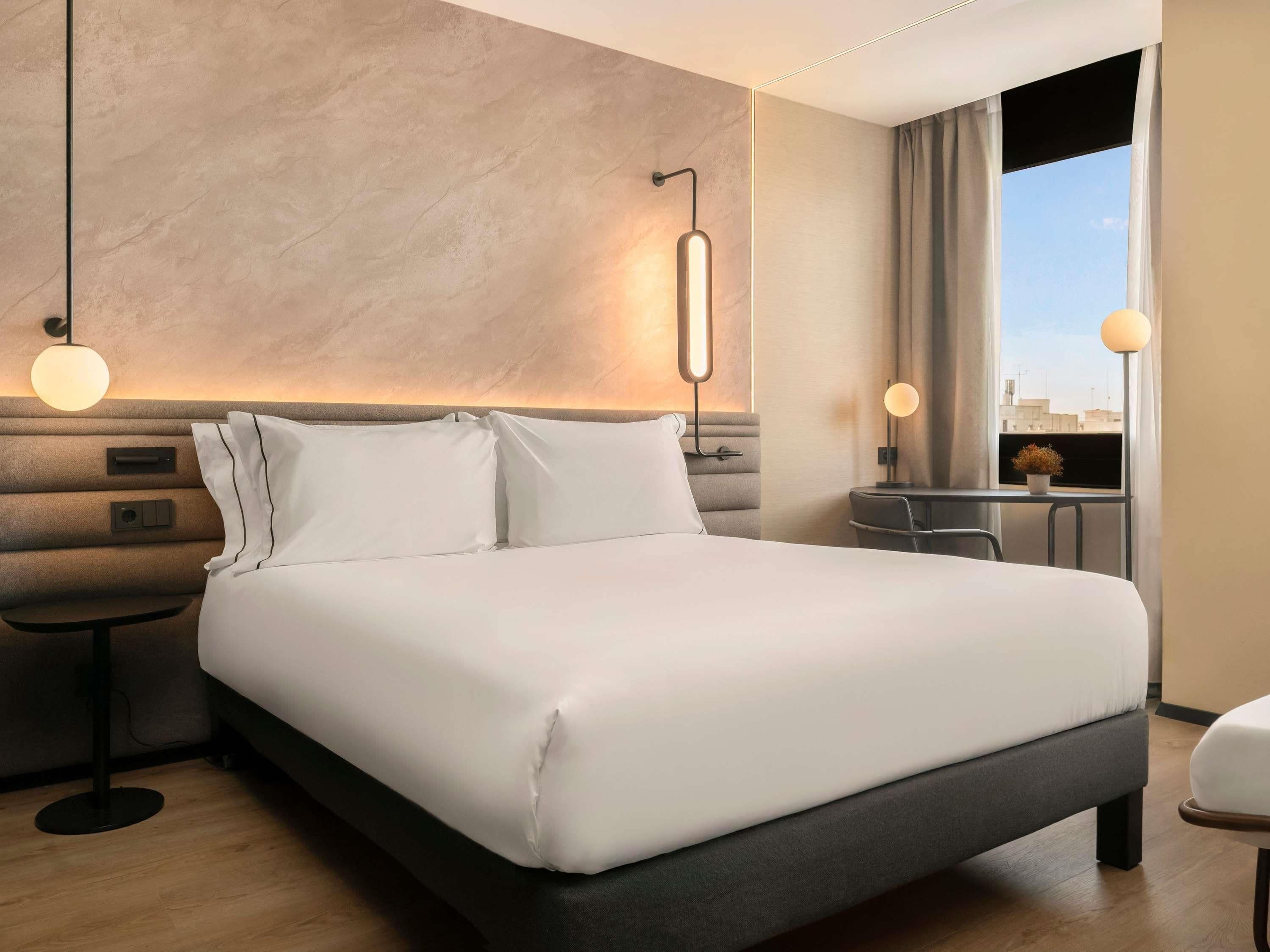 A hotel room with a large bed, bedside tables, a desk, and a view of the city through the window.
