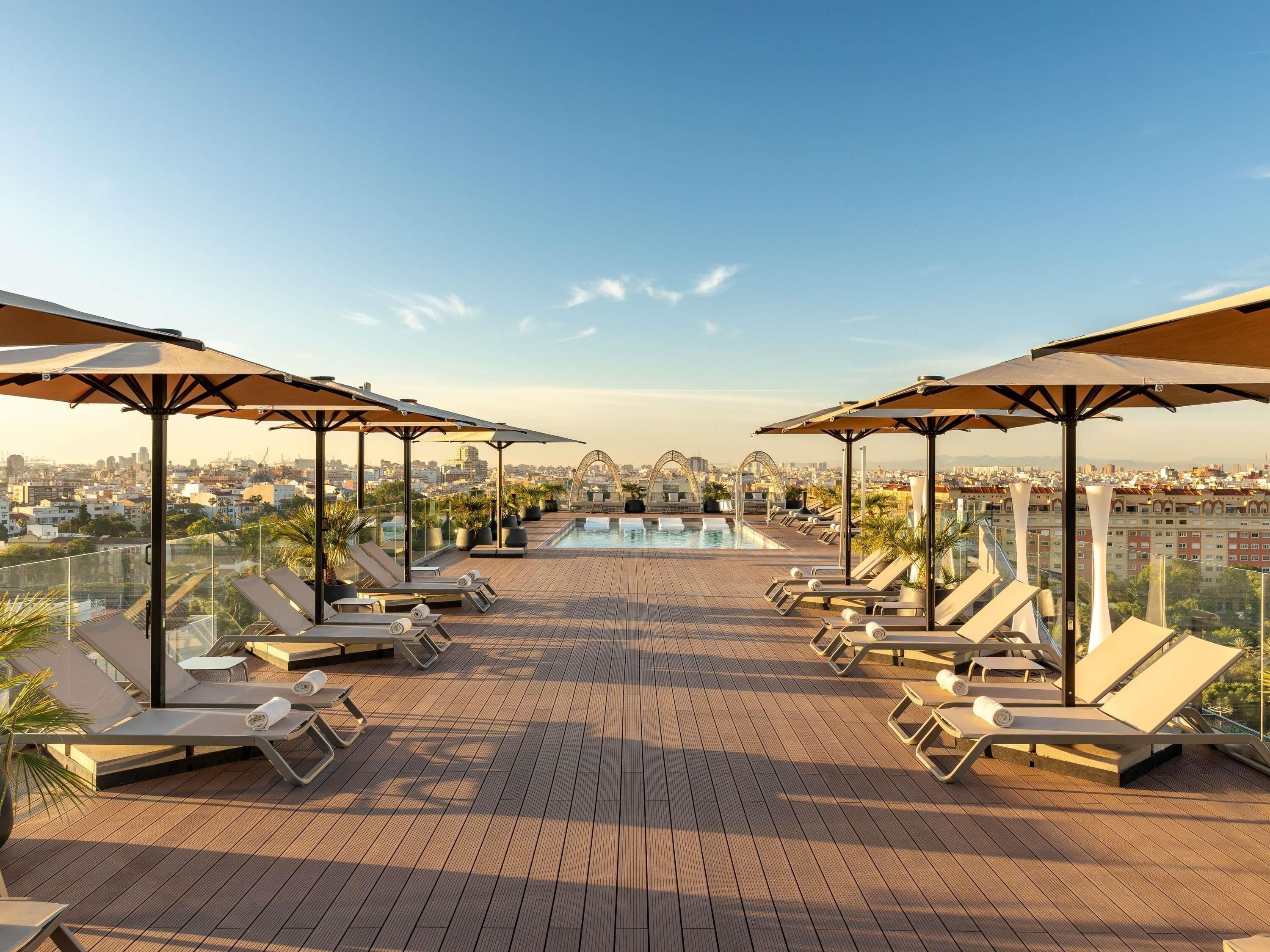 A rooftop pool area with lounge chairs and umbrellas, overlooking a cityscape.