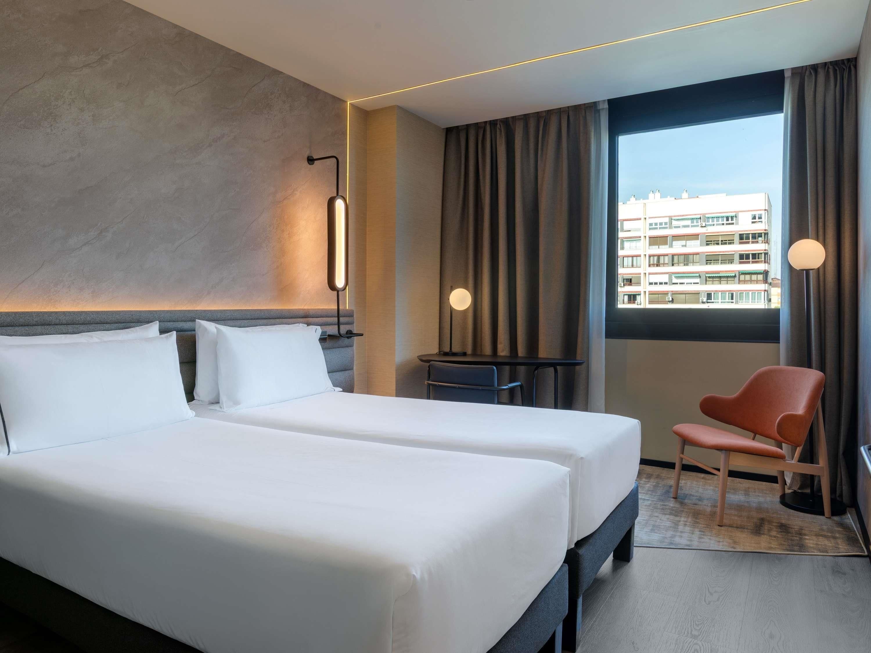 A hotel room with two beds, a desk, a chair, a lamp, and a view of buildings through the window.