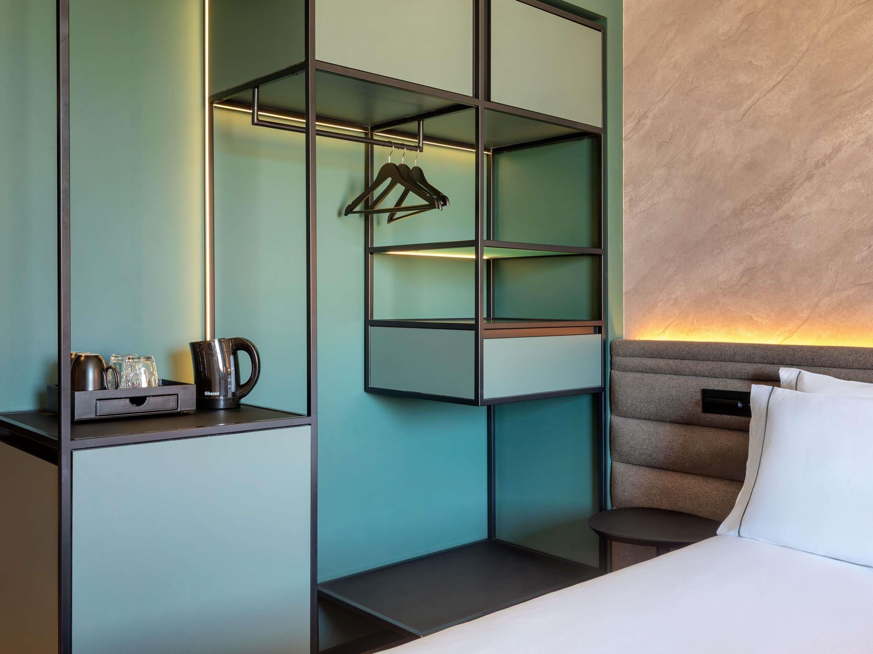 Habitación de hotel con cama, mesita de noche, estante en la pared con tetera y vasos, y perchero.