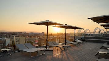 Zona de lounge en la azotea con sillones de sol, sombrillas y vista a la ciudad al atardecer.