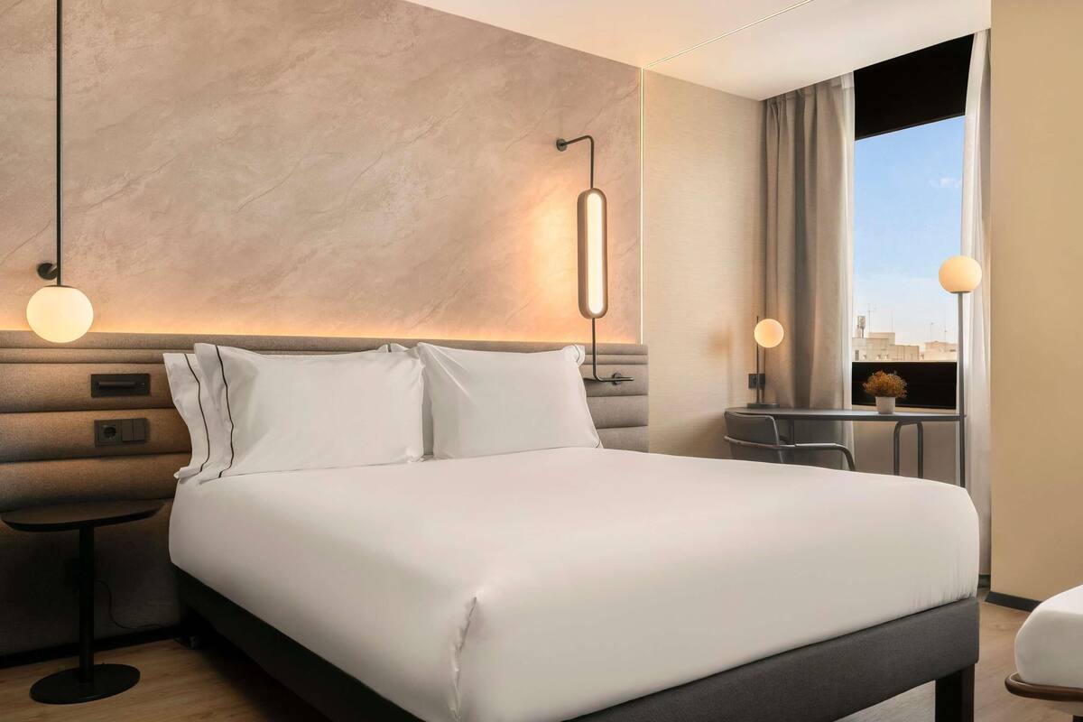 Une chambre d’hôtel avec un grand lit, des tables de chevet, un bureau et une vue sur la ville par la fenêtre.