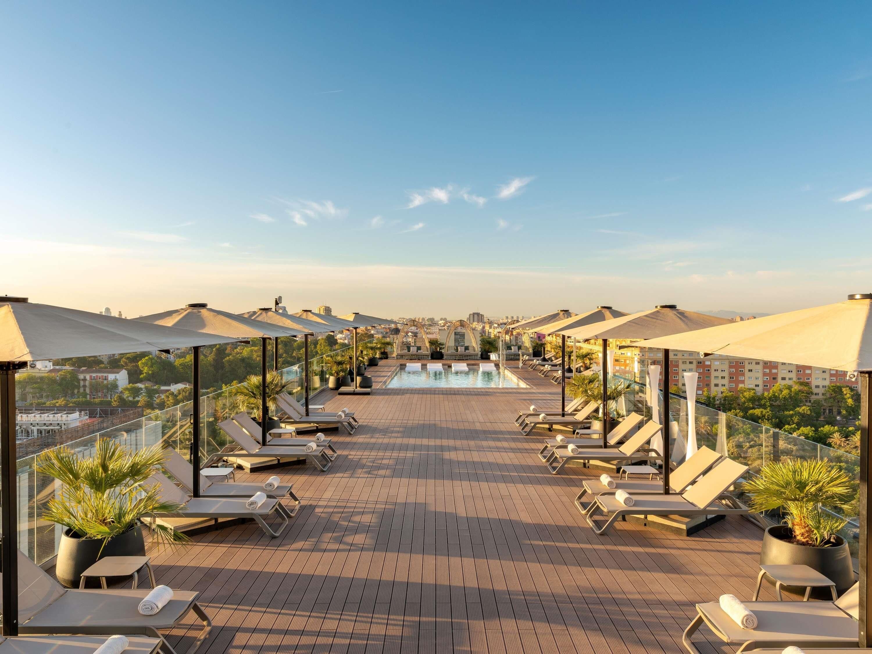 A rooftop pool area with sun loungers and umbrellas, a clear sky, and a cityscape in the background.