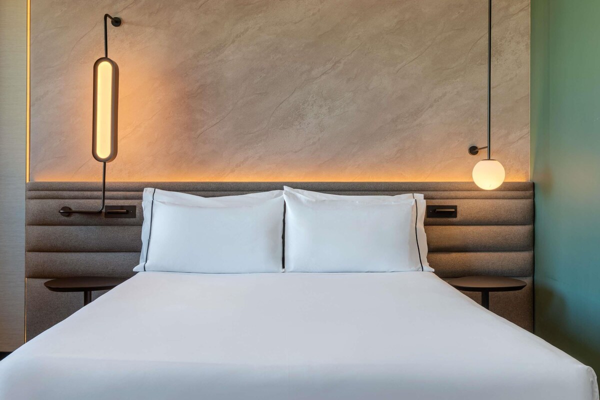 A hotel room with a bed, two bedside tables, and a wall-mounted light fixture.
