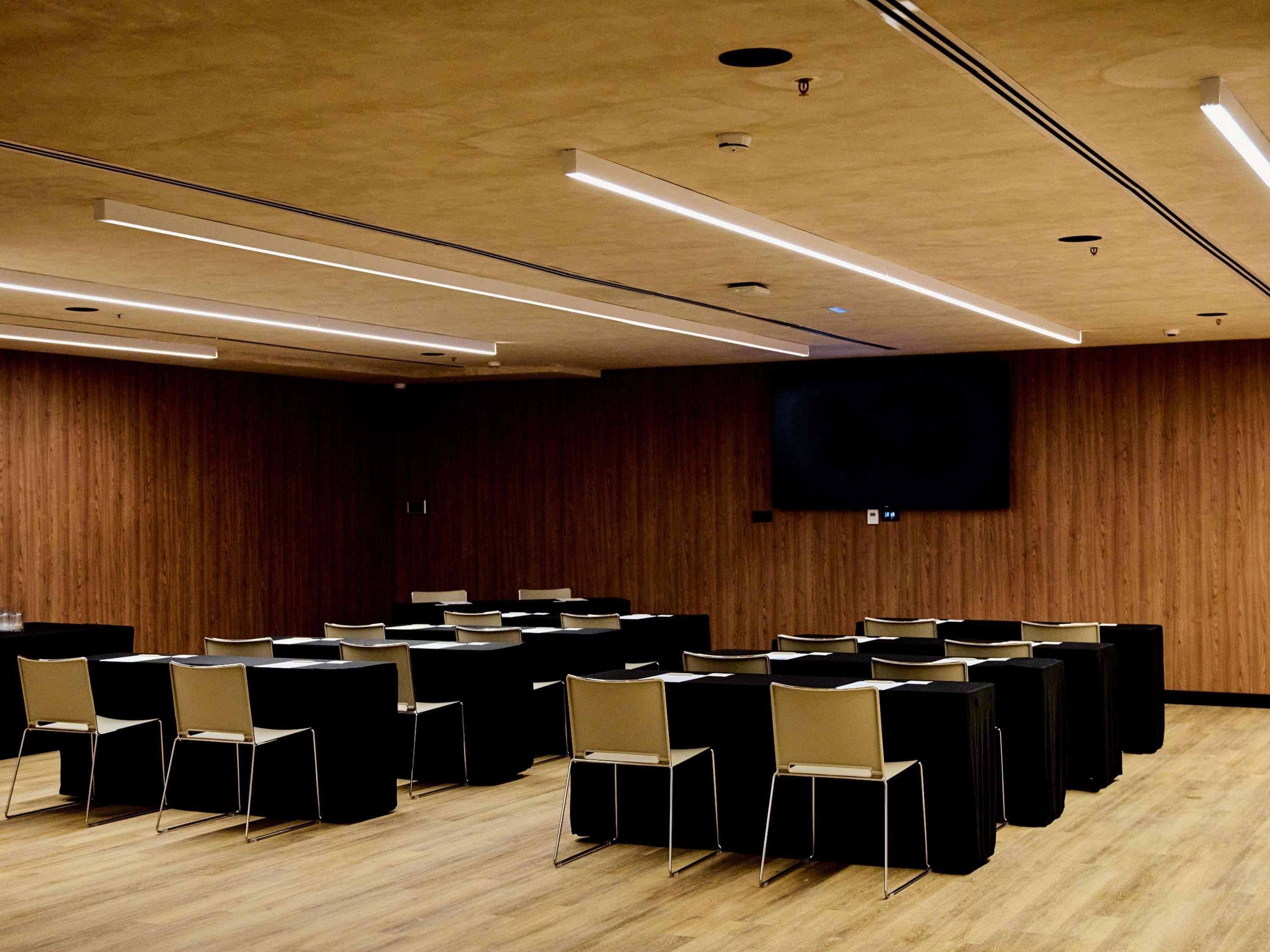 Een vergaderzaal met rijen stoelen, een groot scherm en houten lambrisering.