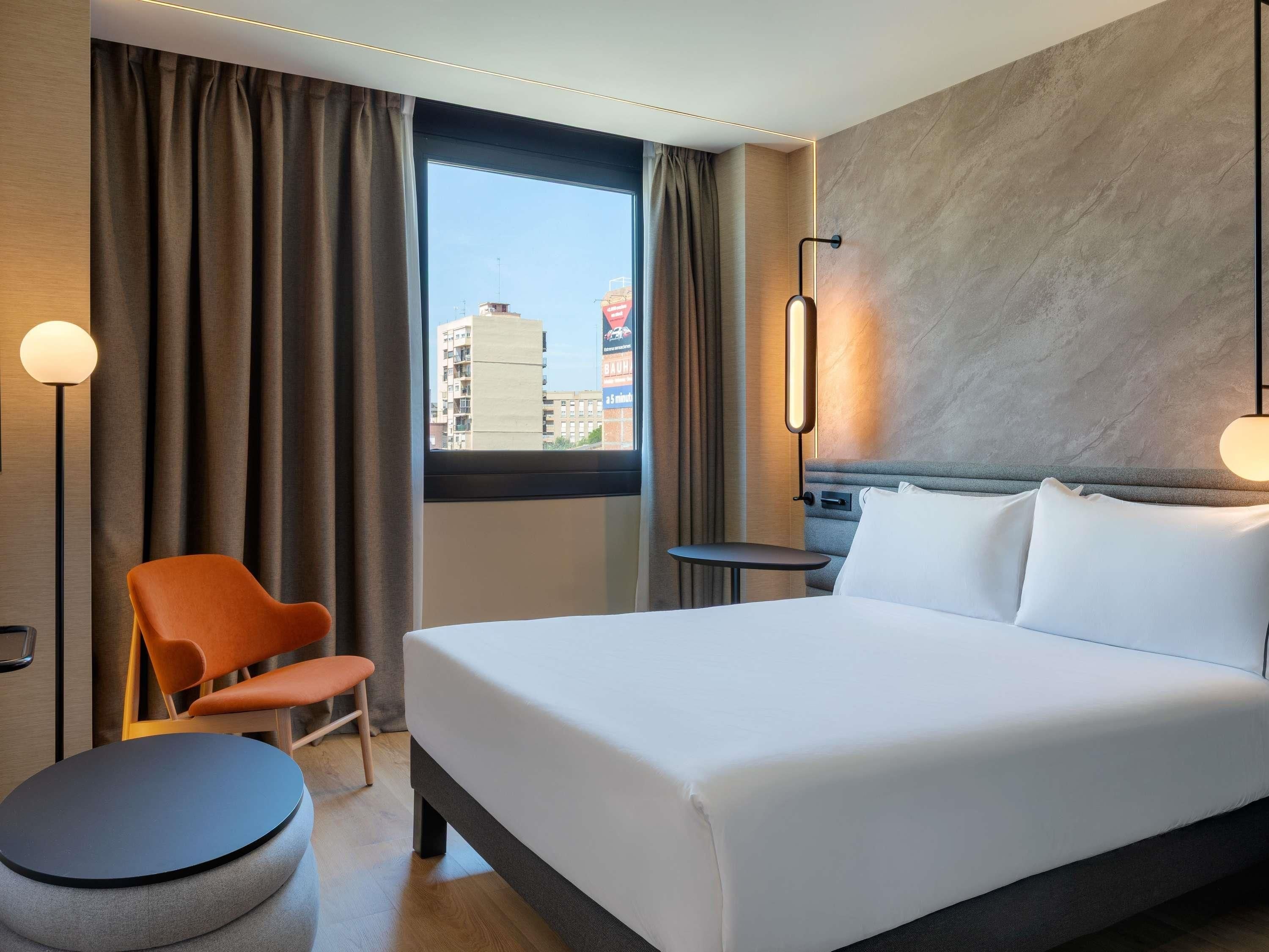 Habitación de hotel con cama, una silla, una mesita, una lámpara y vistas a edificios desde la ventana.