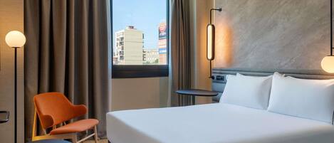 Habitación de hotel con cama, una silla, una mesita, una lámpara y vistas a edificios desde la ventana.