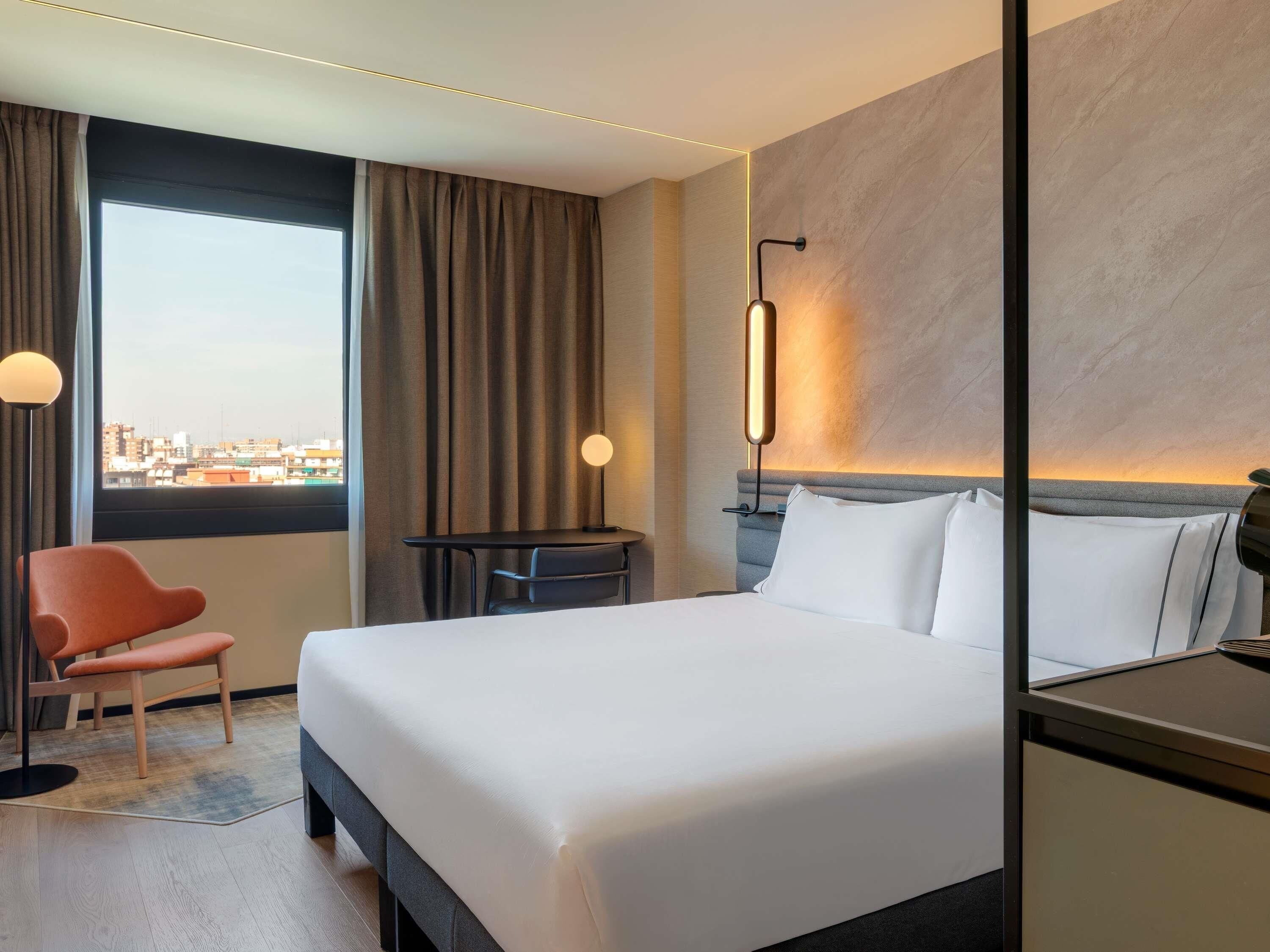 A hotel room with a bed, a desk, a chair, a window with curtains, and a city view.