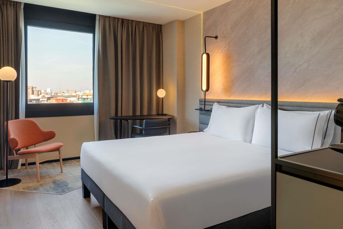 Habitación de hotel con cama, escritorio, silla, ventana con cortinas y vista a la ciudad.