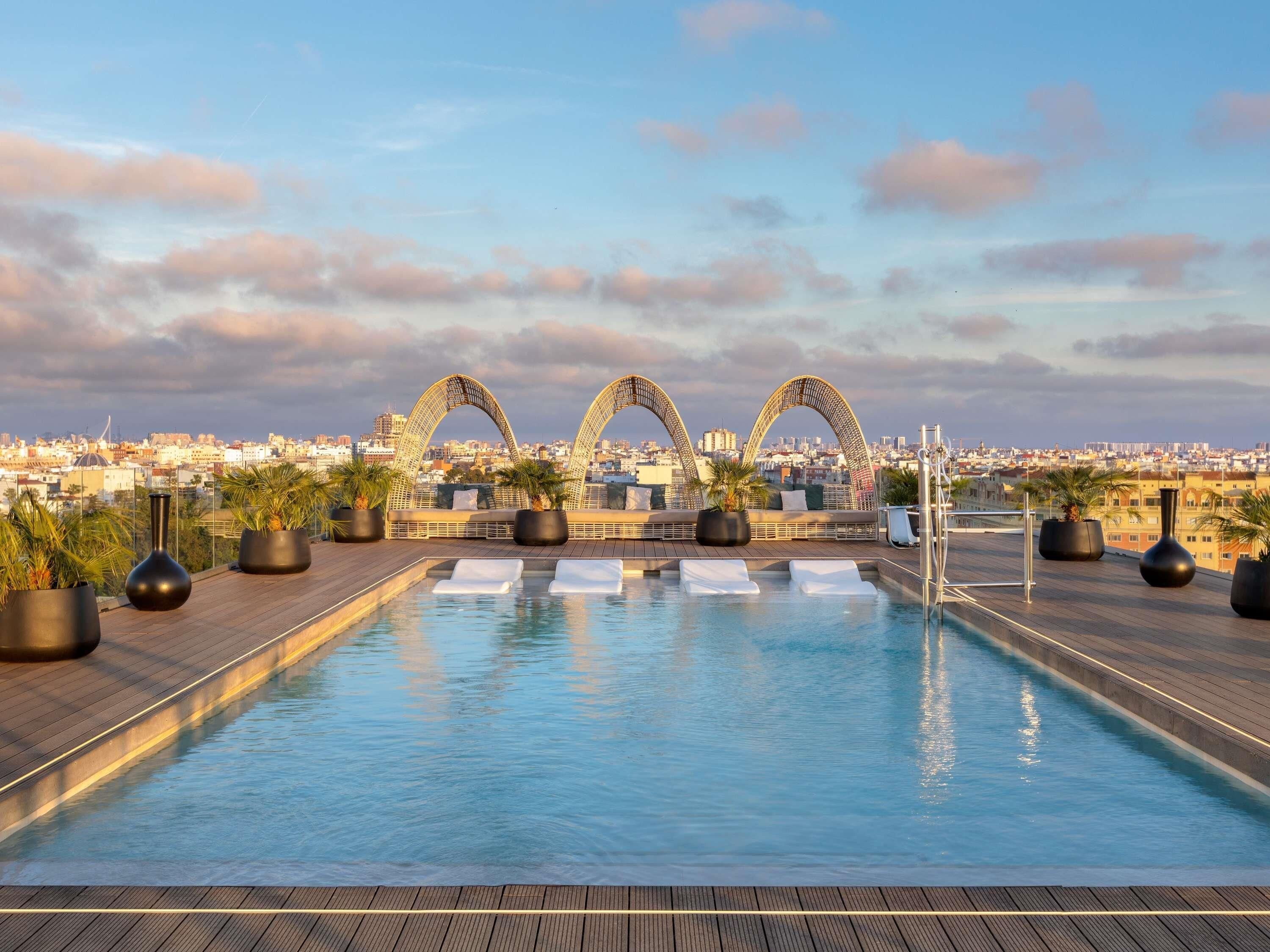Piscina en la azotea con sillones y plantas en macetas, con vistas a un paisaje urbano.