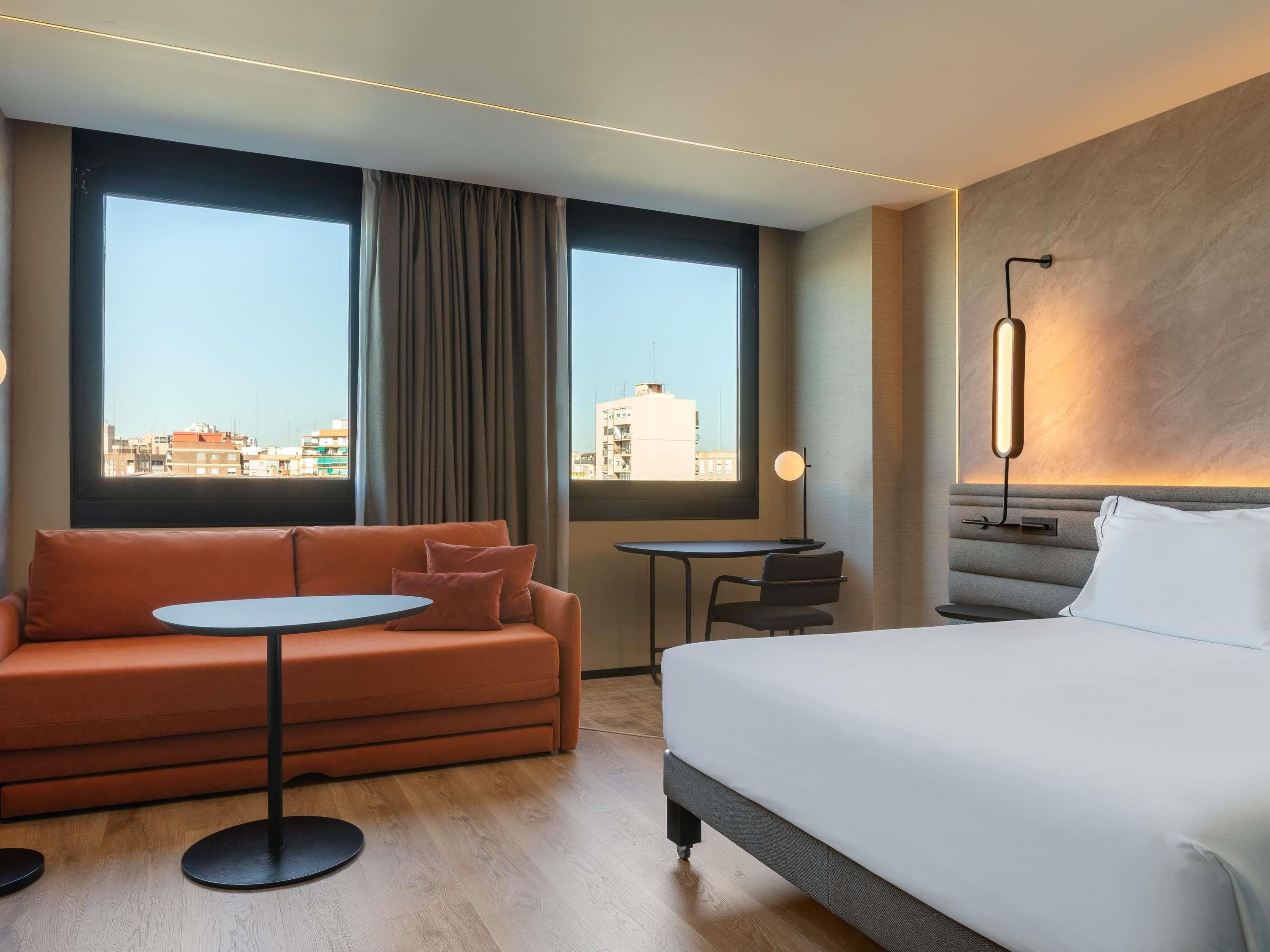 Habitación de hotel con cama, sofá, una mesita y vistas de la ciudad a través de amplias ventanas.