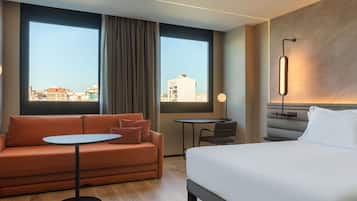 Habitación de hotel con cama, sofá, una mesita y vistas de la ciudad a través de amplias ventanas.