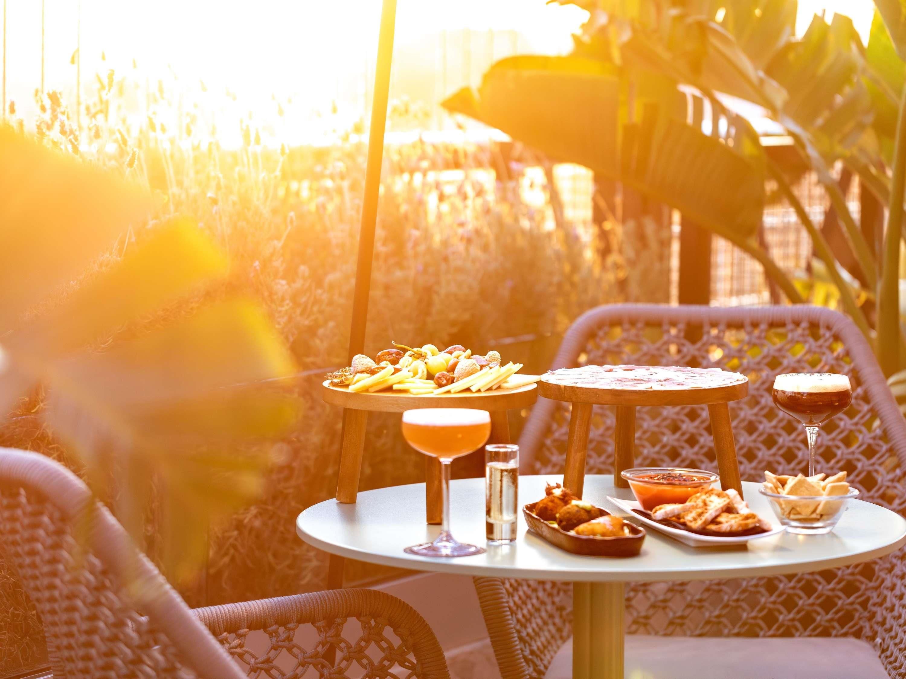 Una mesa con comida y bebidas, rodeada de sillas de mimbre, en un entorno exterior soleado.