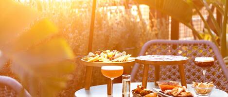 Una mesa con comida y bebidas, rodeada de sillas de mimbre, en un entorno exterior soleado.