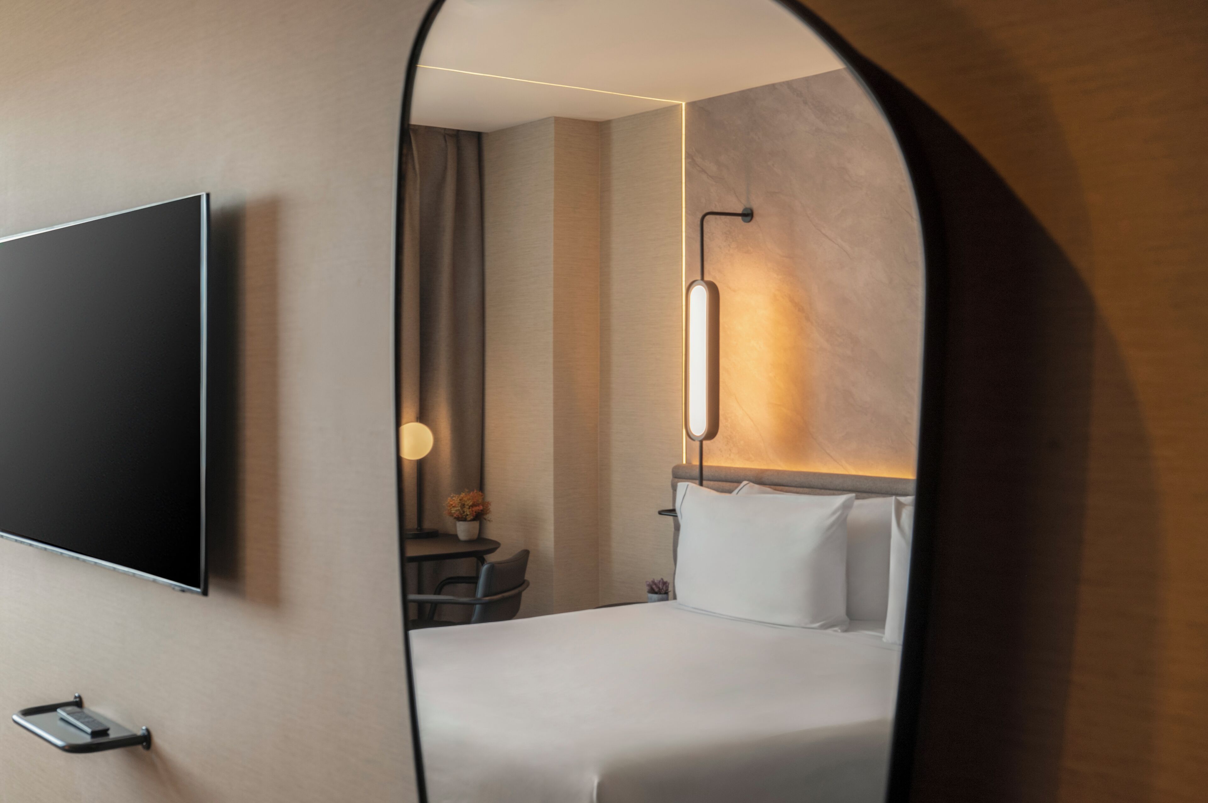 Habitación de hotel con cama, televisor montado en la pared, mesita de noche con lámpara y espejo que refleja la habitación.