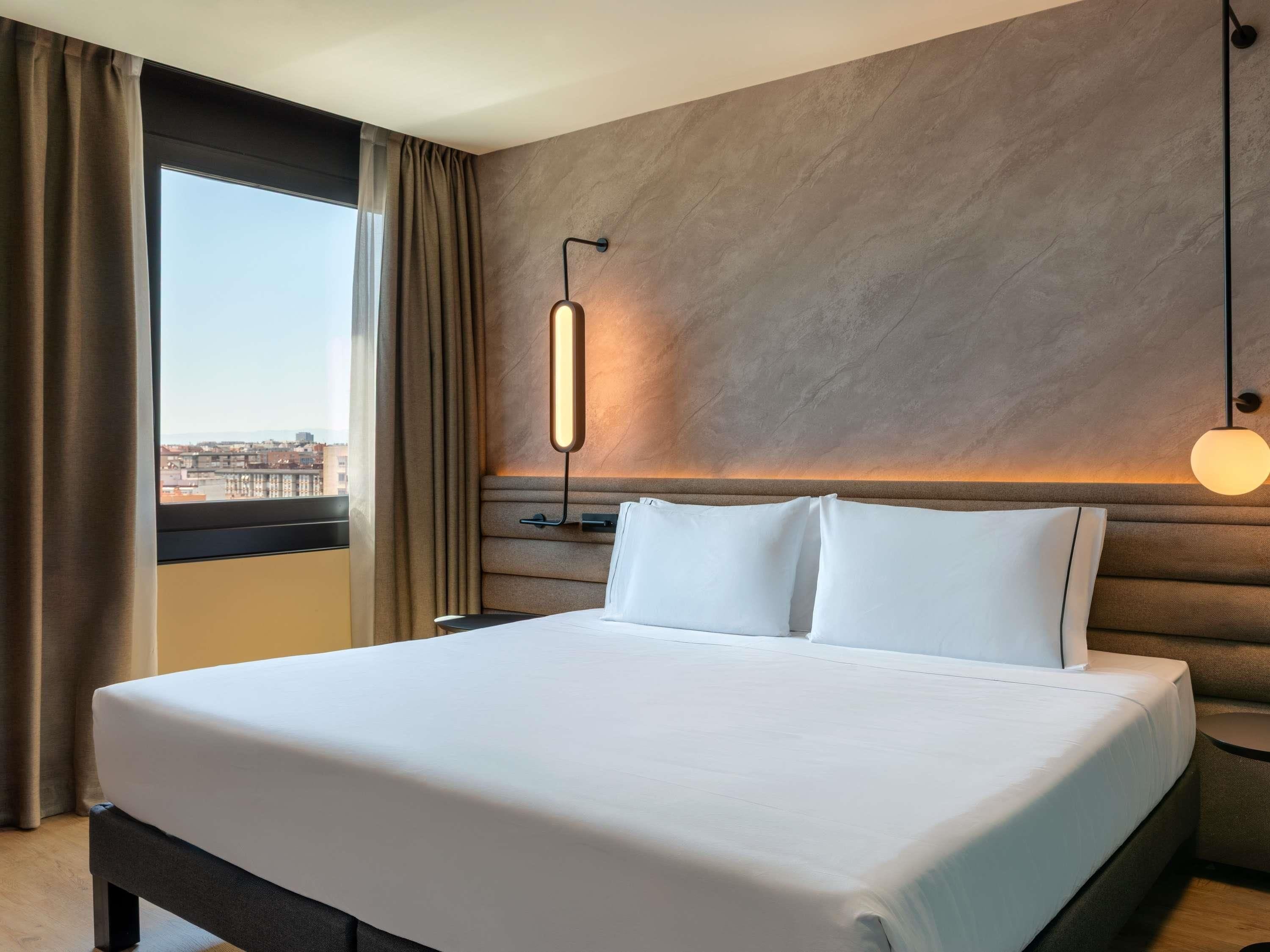 Une chambre d’hôtel avec un grand lit, une tête de lit dotée d’un luminaire, une vue sur la ville par la fenêtre et une table de chevet avec une lampe.