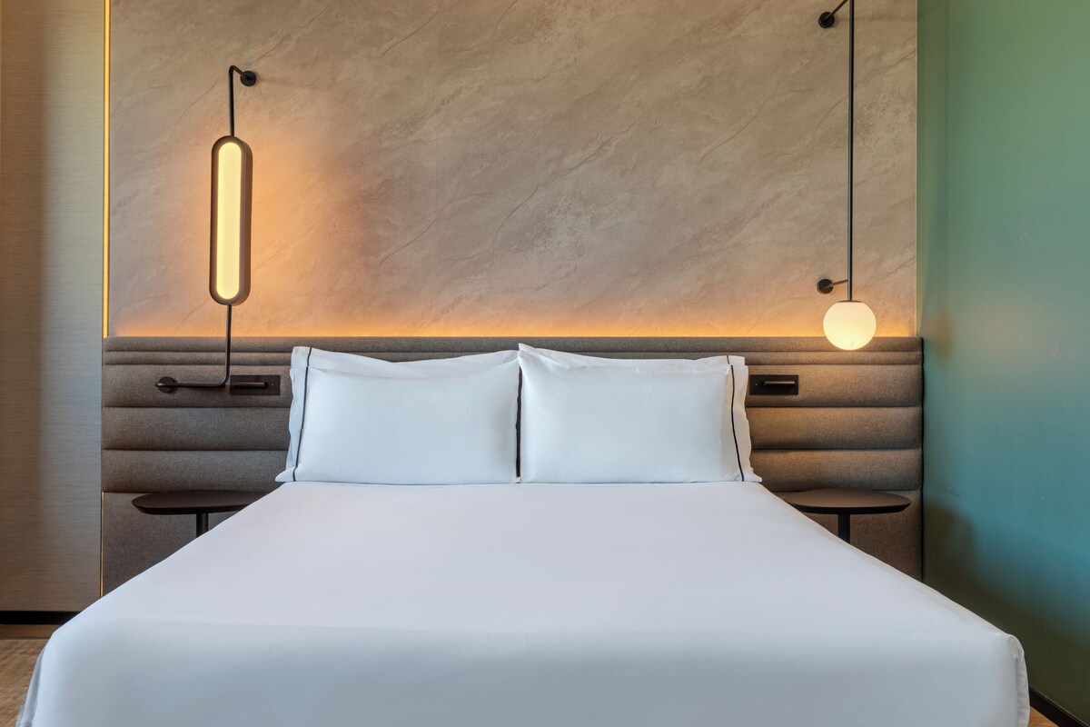 Una cama bien hecha con cabecera, dos mesitas de noche e iluminación en la pared.