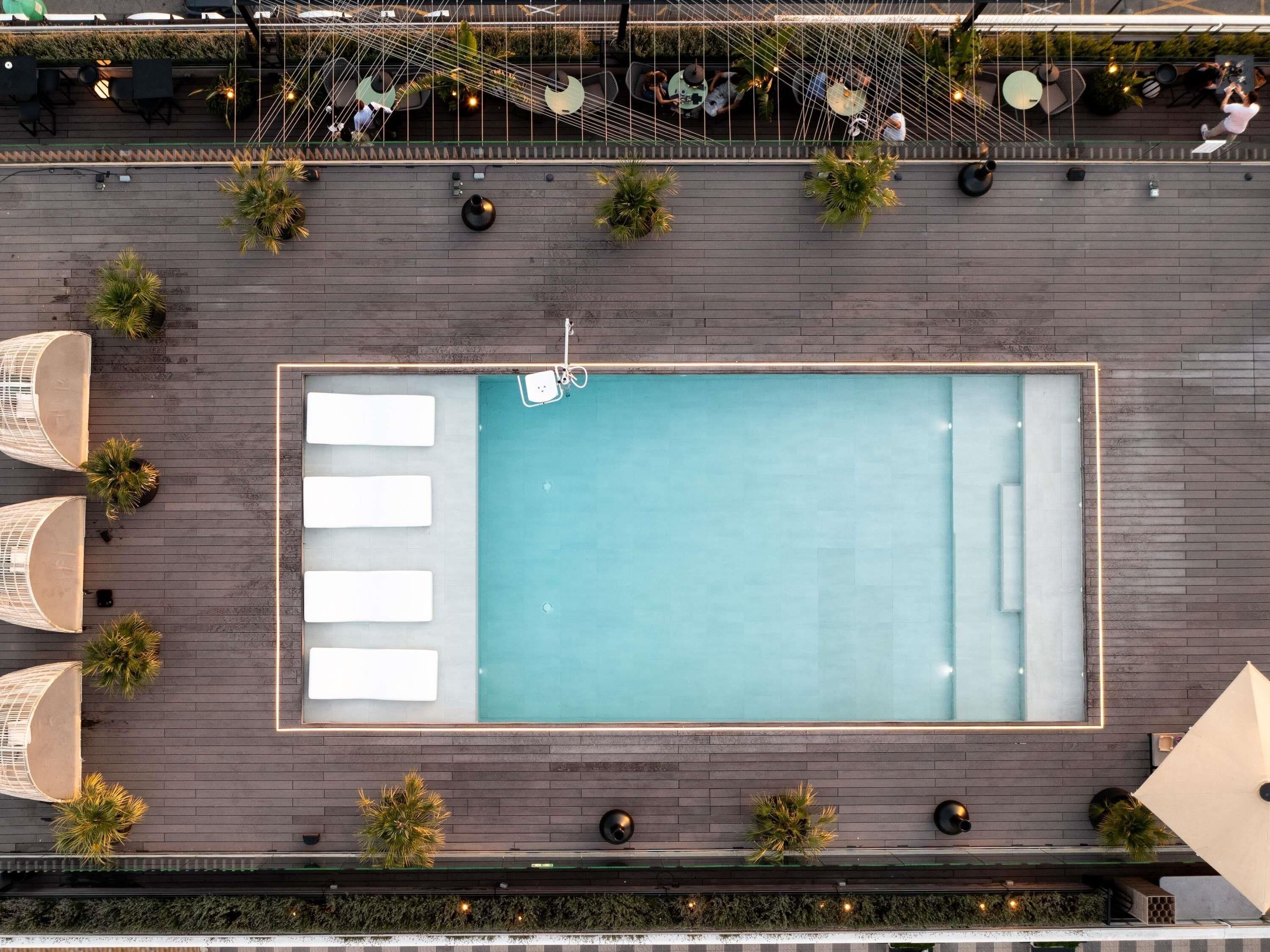 An aerial view of a rooftop pool area with lounge chairs, a pool, and surrounding greenery.