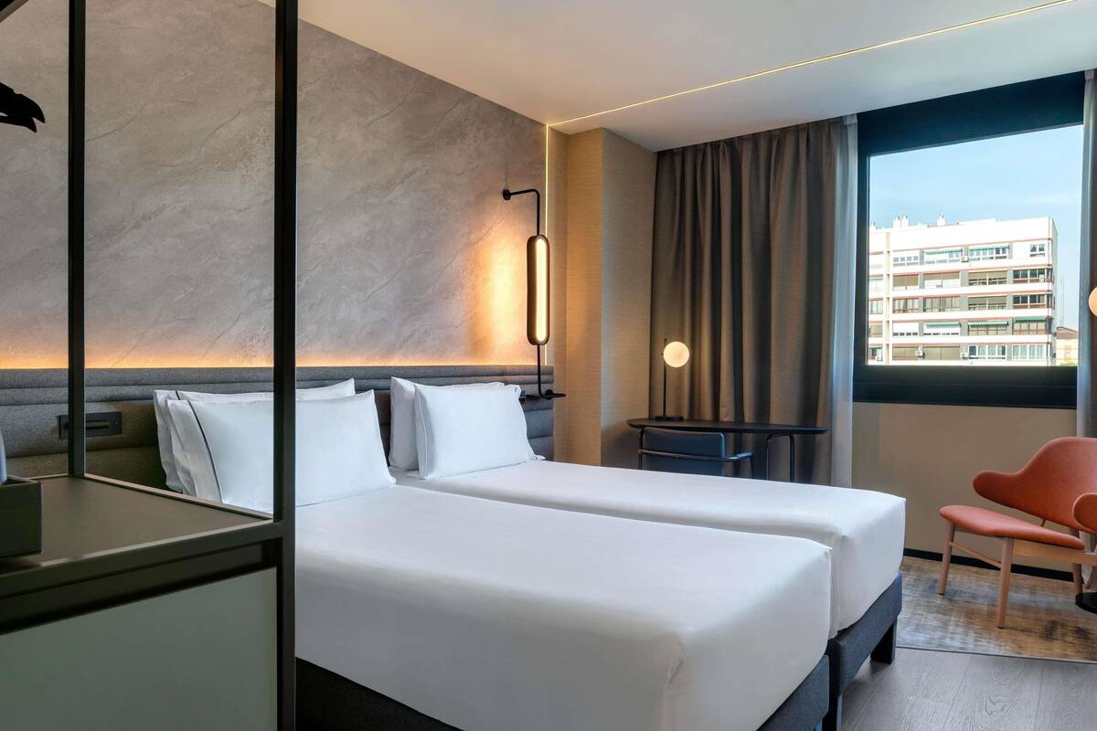Une chambre d’hôtel avec deux lits, un bureau, une chaise, une fenêtre donnant sur des immeubles et une lampe fixée au mur.