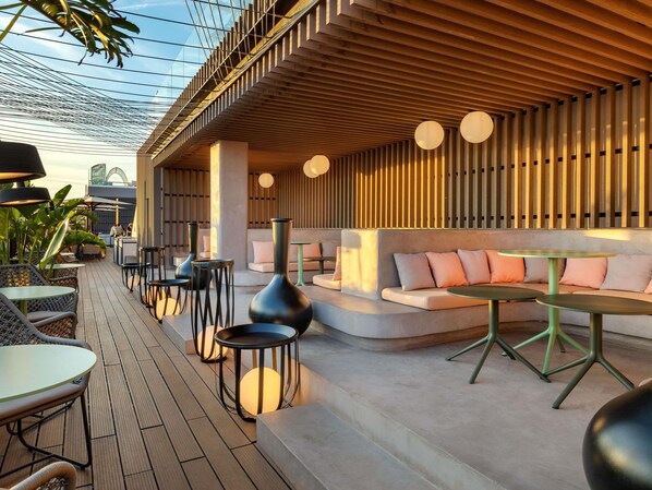Un área de estar al aire libre con mesas redondas, asientos acolchados, techo de listones de madera y una estructura de vidrio superior.