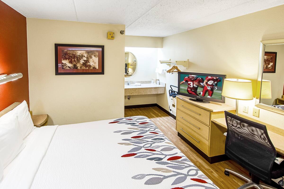Ein Hotelzimmer mit einem Bett, einem Schreibtisch mit Stuhl, einem Fernseher, einem Spiegel und einem Badezimmer im Hintergrund.