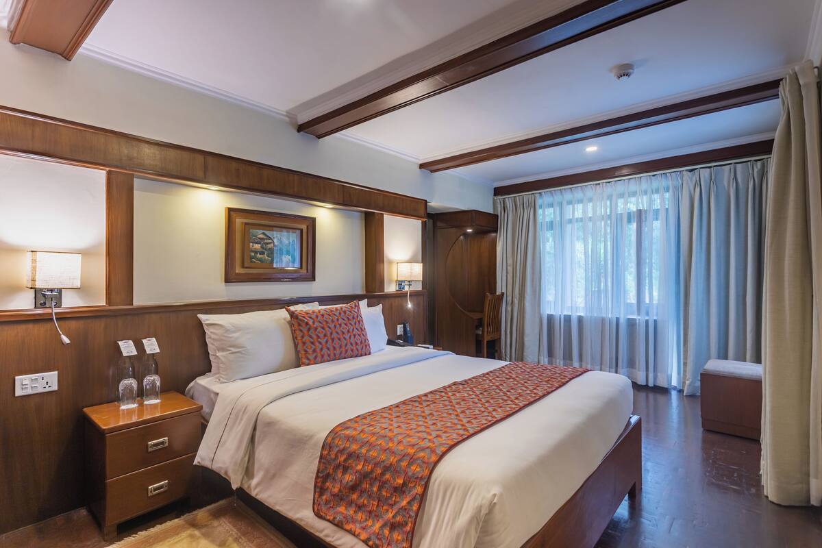 A hotel room with a large bed, wooden headboard, bedside lamps, a nightstand, a window with curtains, and a small seating area.