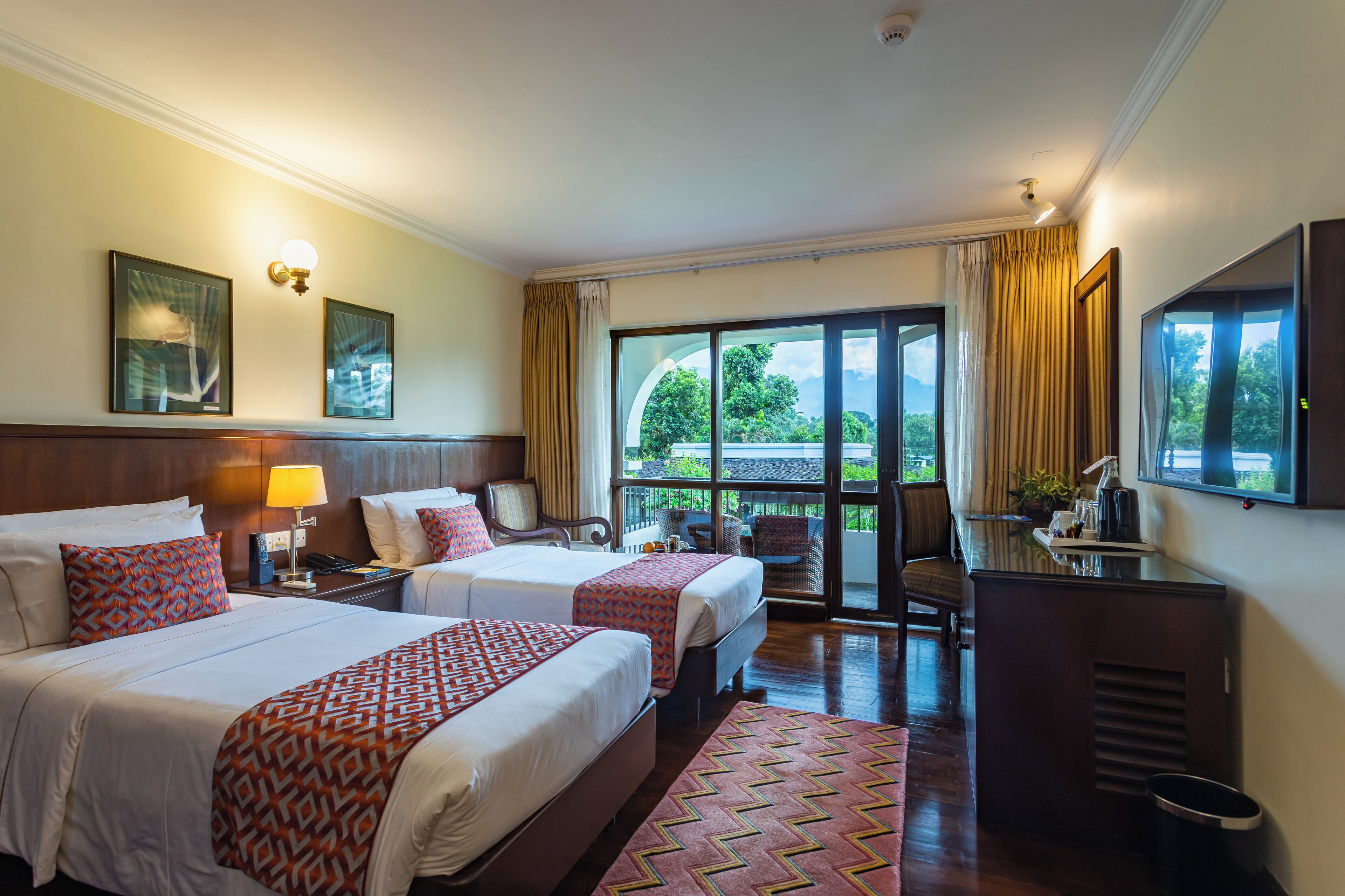 A hotel room with two beds, a desk, a TV, and a balcony with a view of greenery.