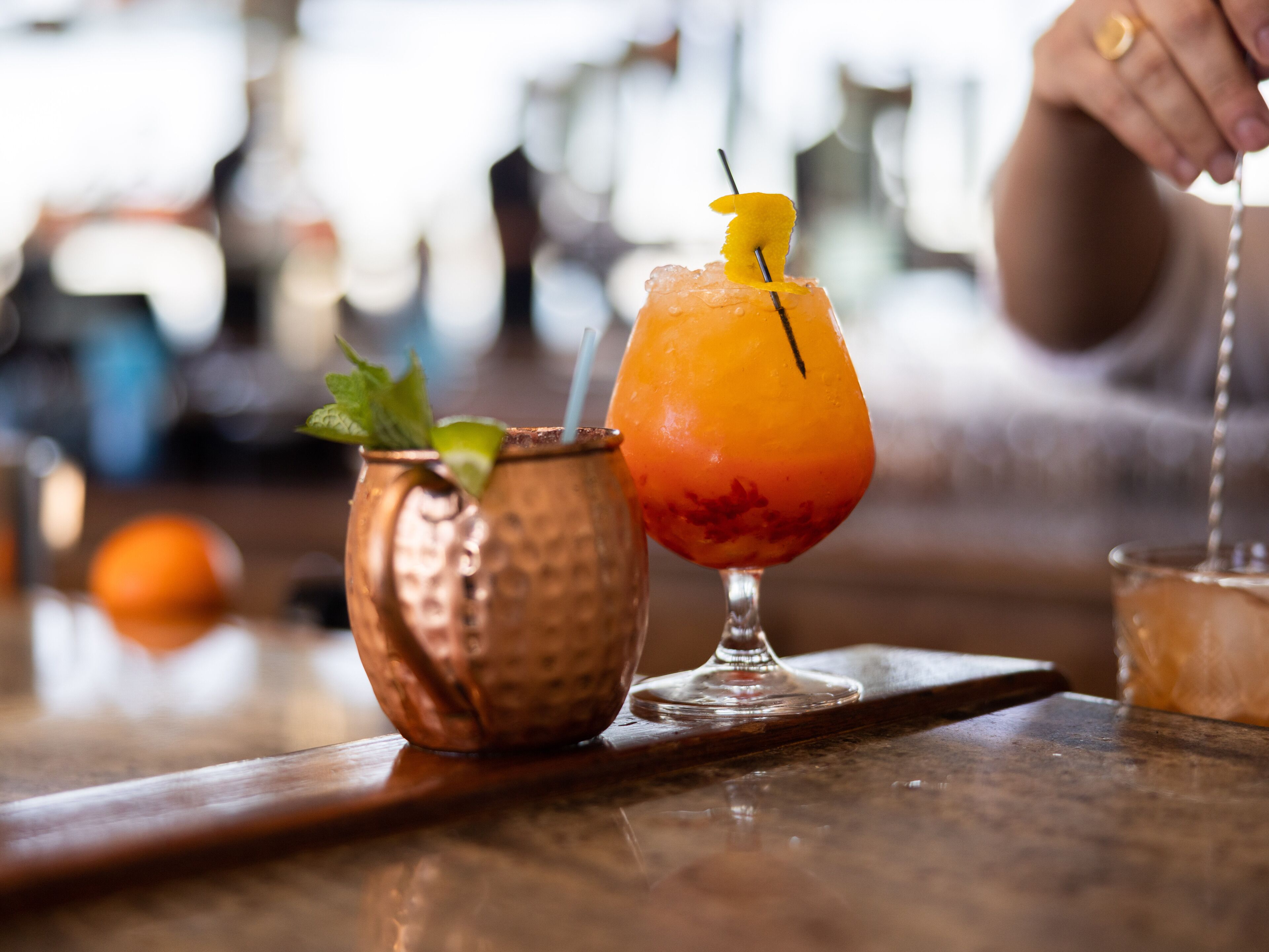 Two cocktails on a bar counter, one in a copper mug and one in a glass with a straw.