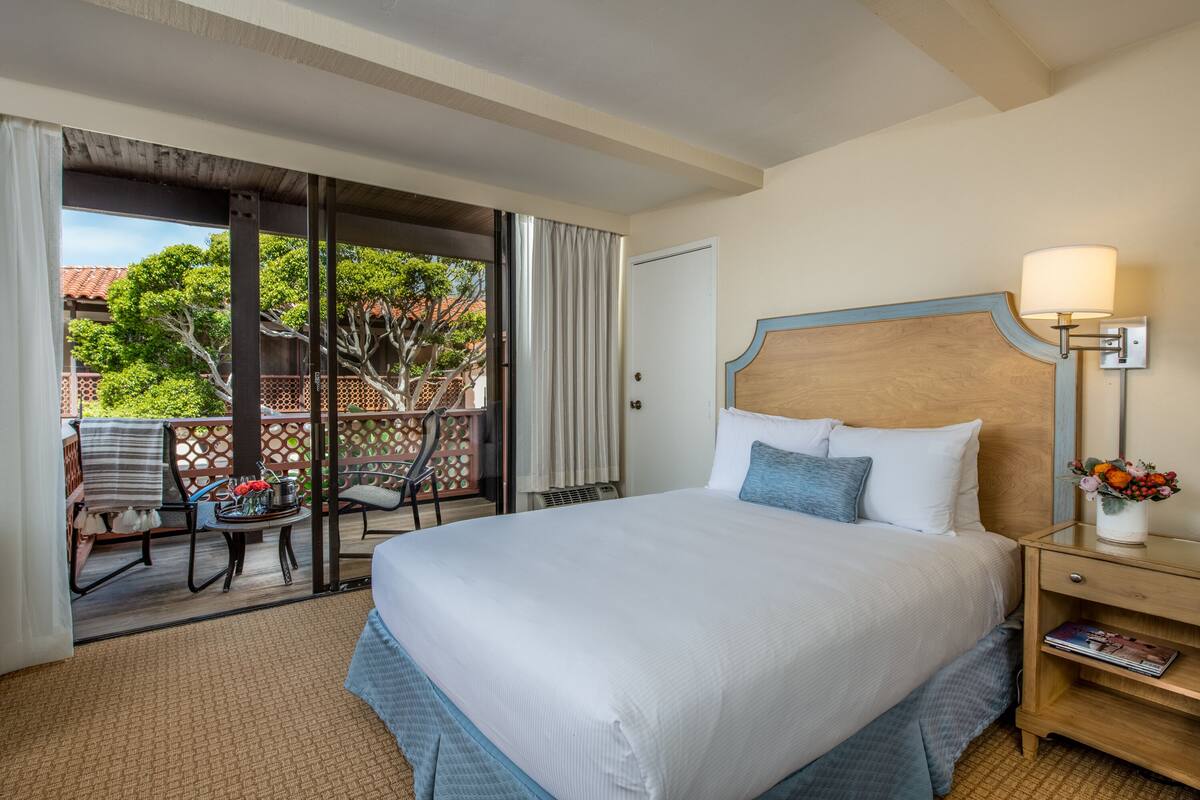 A hotel room with a large bed, a bedside table with a lamp, a chair, and a view of the outdoor area.