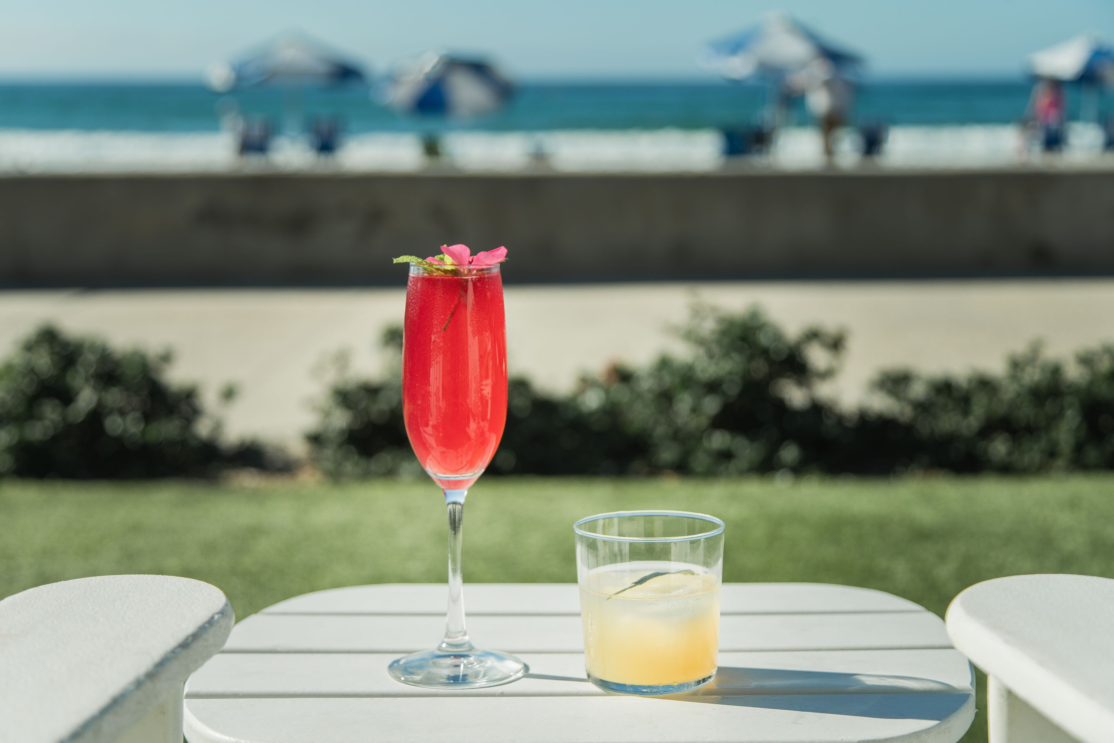 Una mesa con dos bebidas, una en un vaso alto y otra en uno bajo, al aire libre con una playa y sombrillas de fondo.