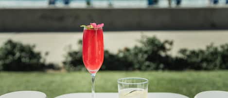 Una mesa con dos bebidas, una en un vaso alto y otra en uno bajo, al aire libre con una playa y sombrillas de fondo.