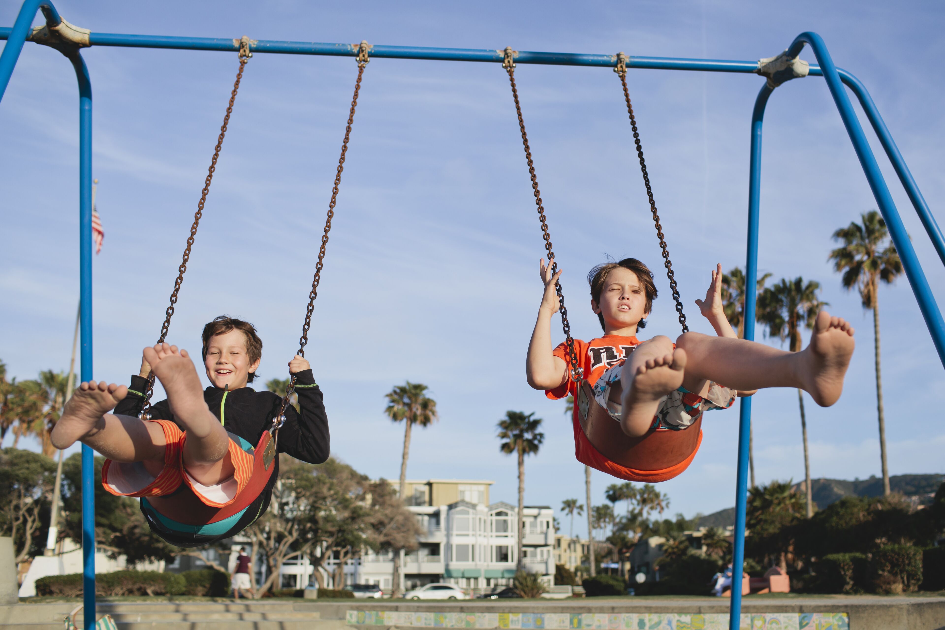 Deux enfants sur des balançoires dans un parc, avec des palmiers et des bâtiments en arrière-plan.