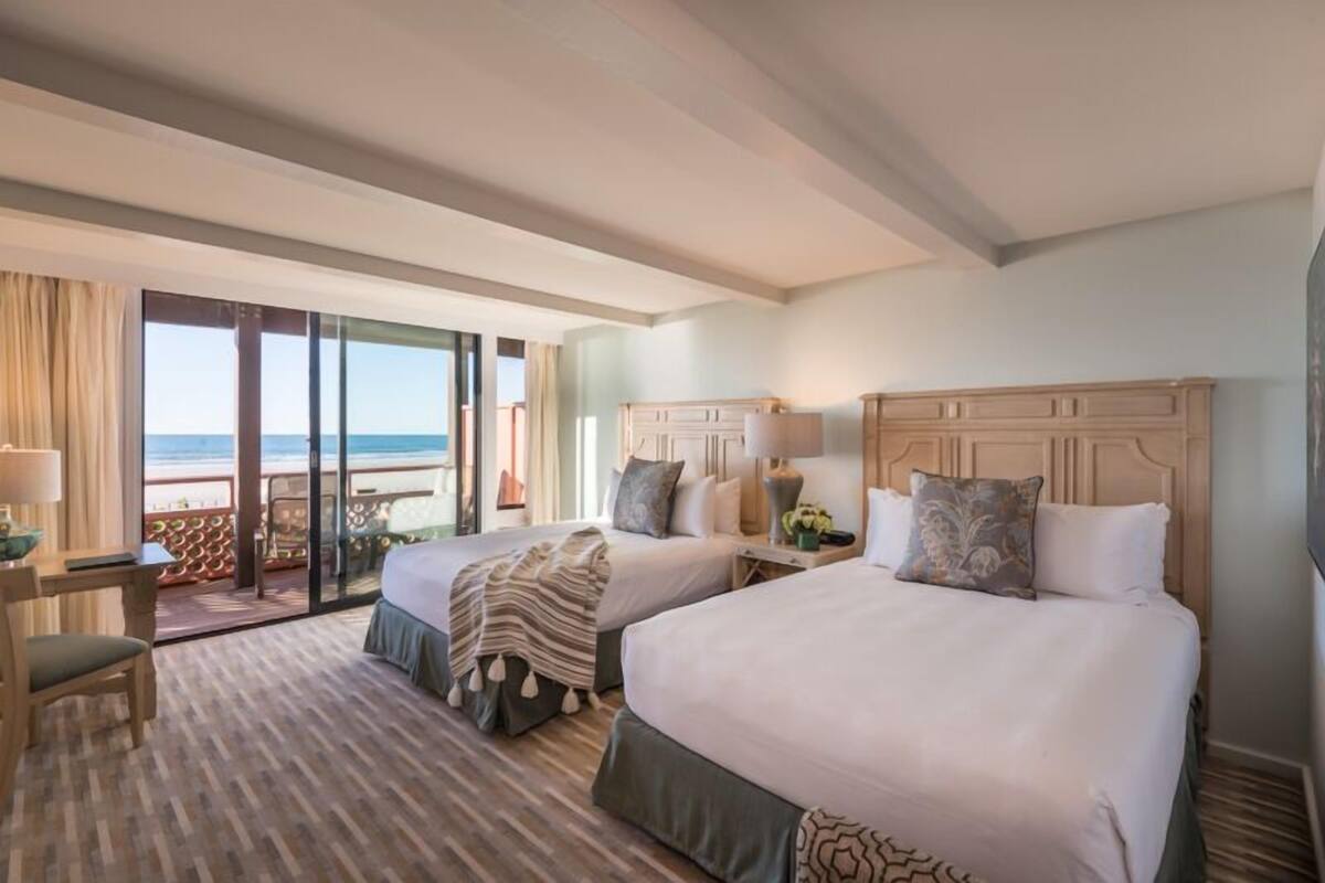 A hotel room with two beds, a desk, and a view of the beach.