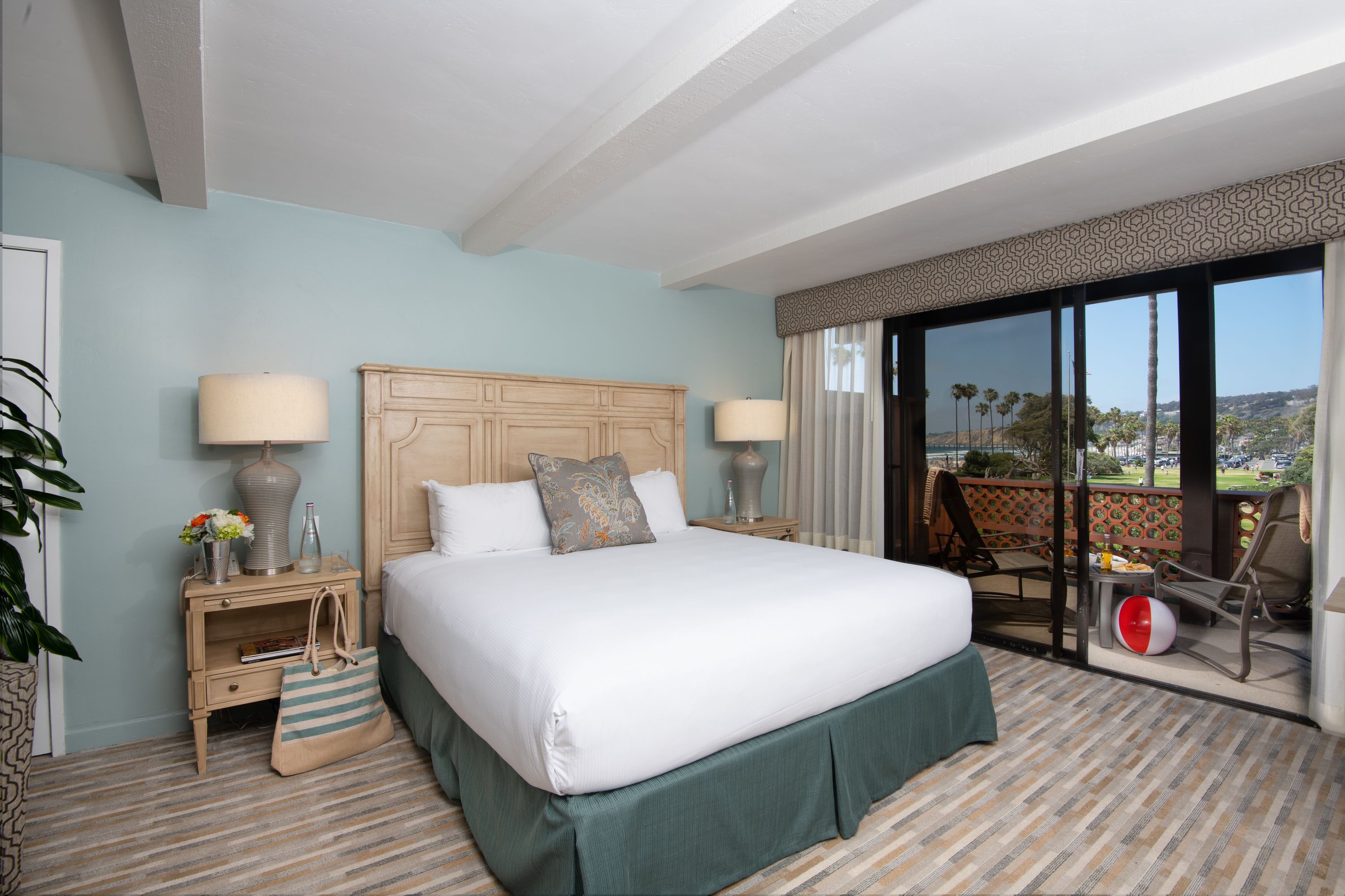 A hotel room with a large bed, two bedside tables with lamps, a nightstand with a lamp and a bag, and a view of the outdoors through a sliding glass door.