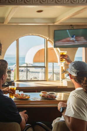 A bar area with a view of the beach, a television screen, and a table with food and drinks.