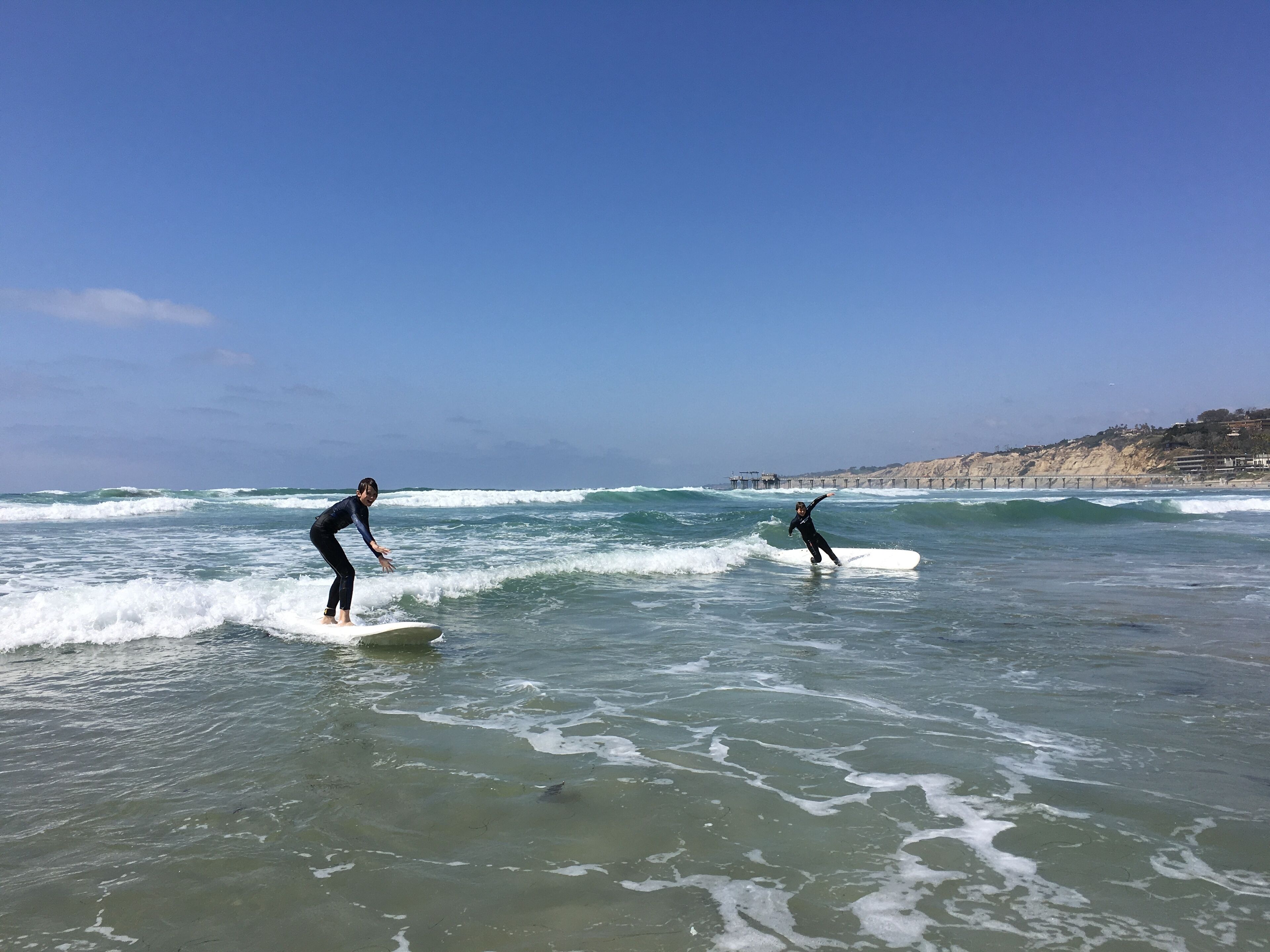 Two surfers on a beach with waves, a pier in the background, and a hilly landscape.