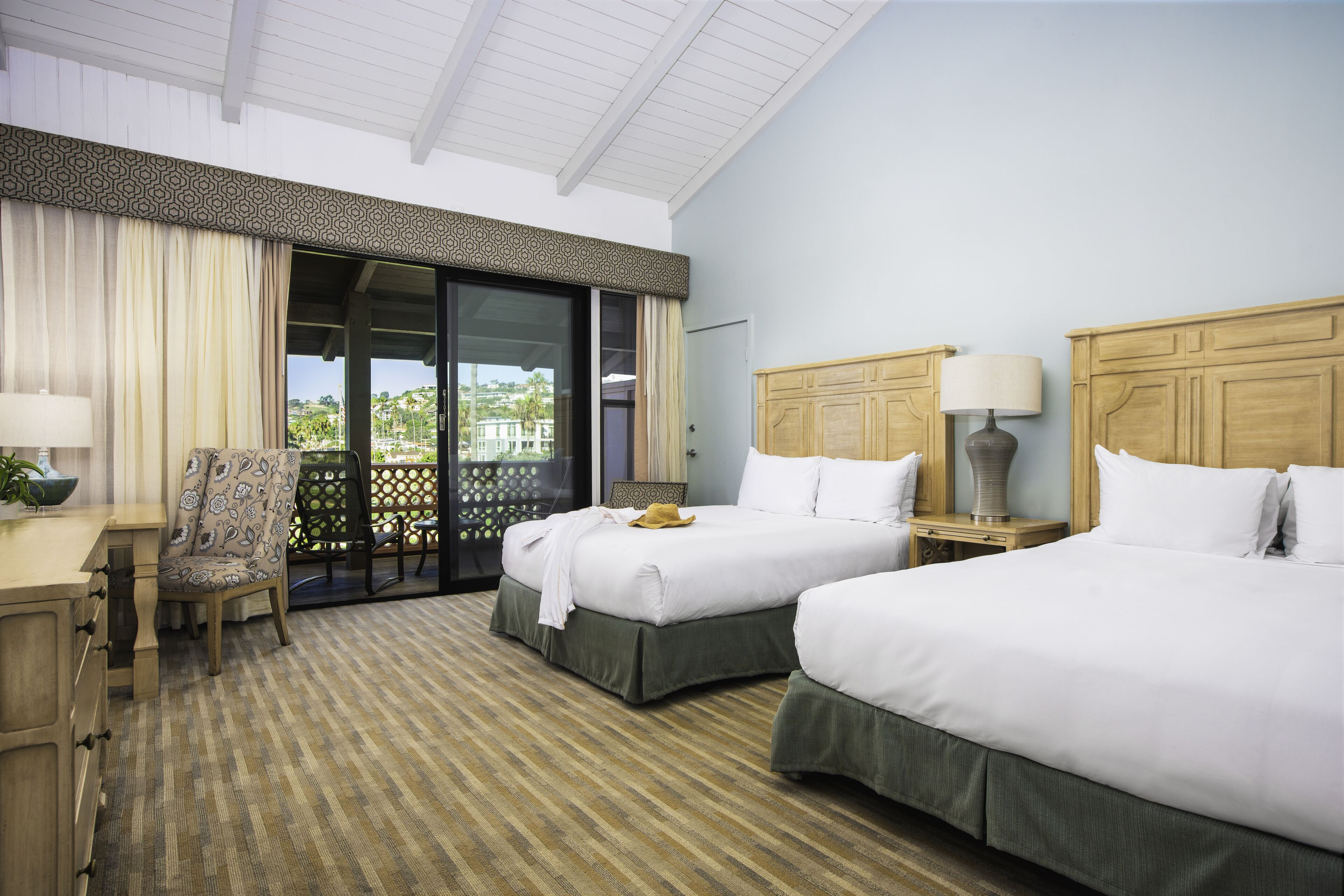 A hotel room with two beds, a desk, a chair, and a view of the outdoors.