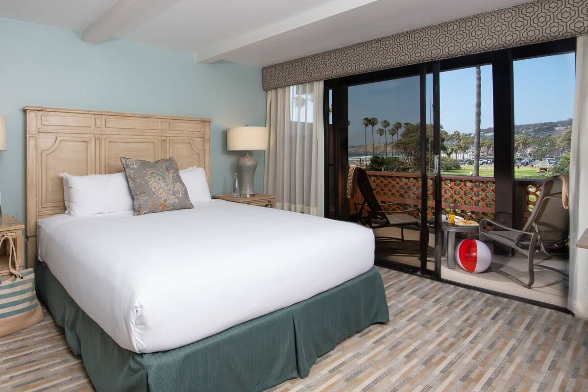 A hotel room with a large bed, two bedside tables with lamps, a chair, a small table with a drink, and a view of palm trees and a distant landscape.
