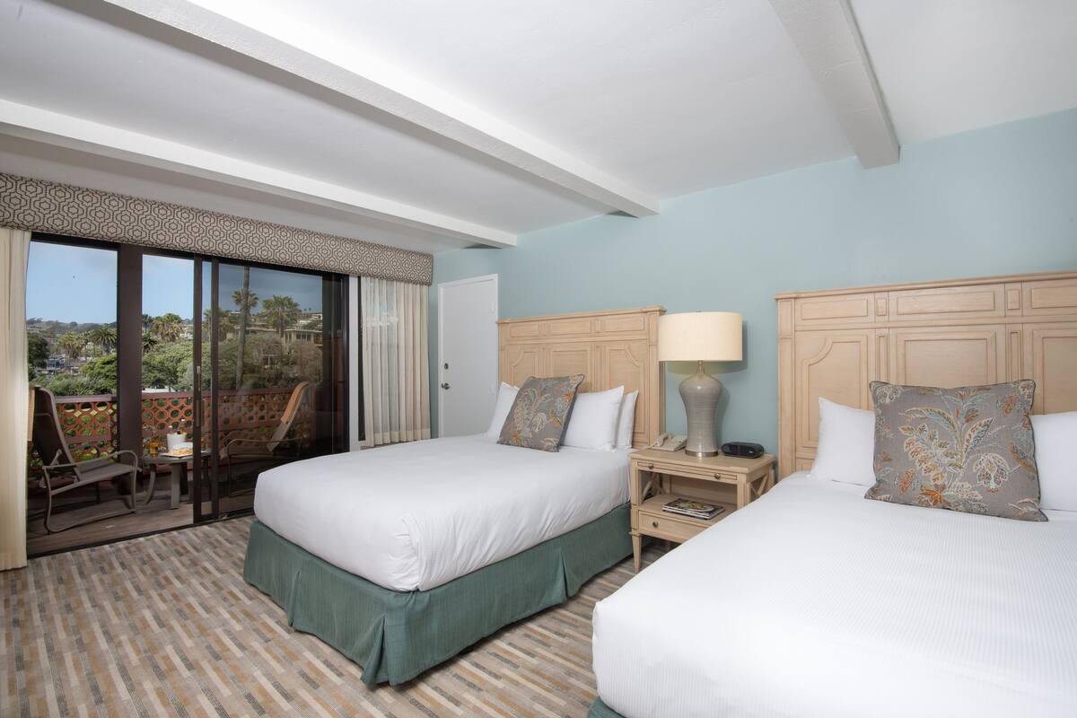 A hotel room with two beds, a balcony with chairs, and a view of palm trees.