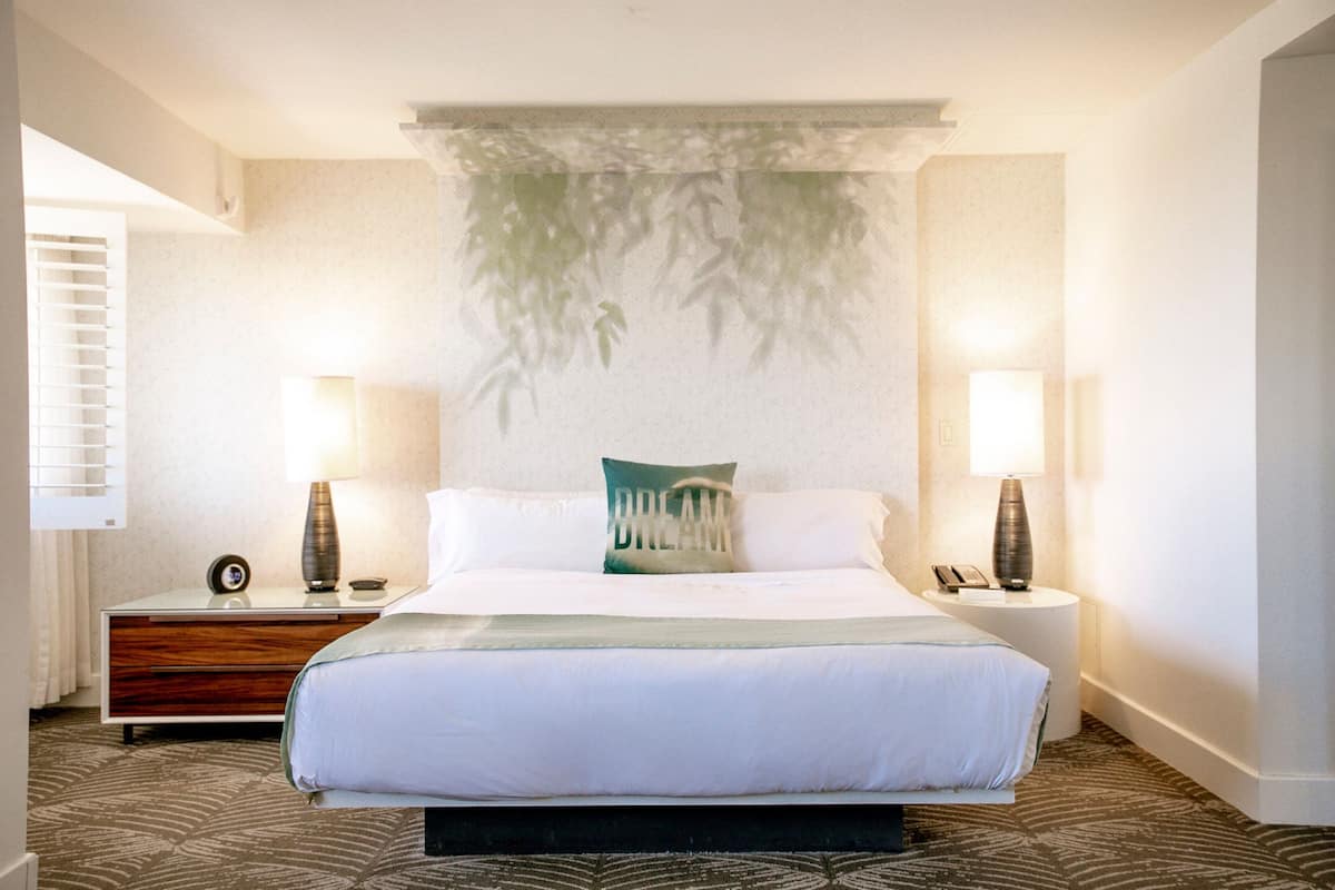 Une chambre d’hôtel avec un grand lit, deux tables de chevet avec des lampes, une œuvre d’art accrochée au mur et une moquette à motifs.