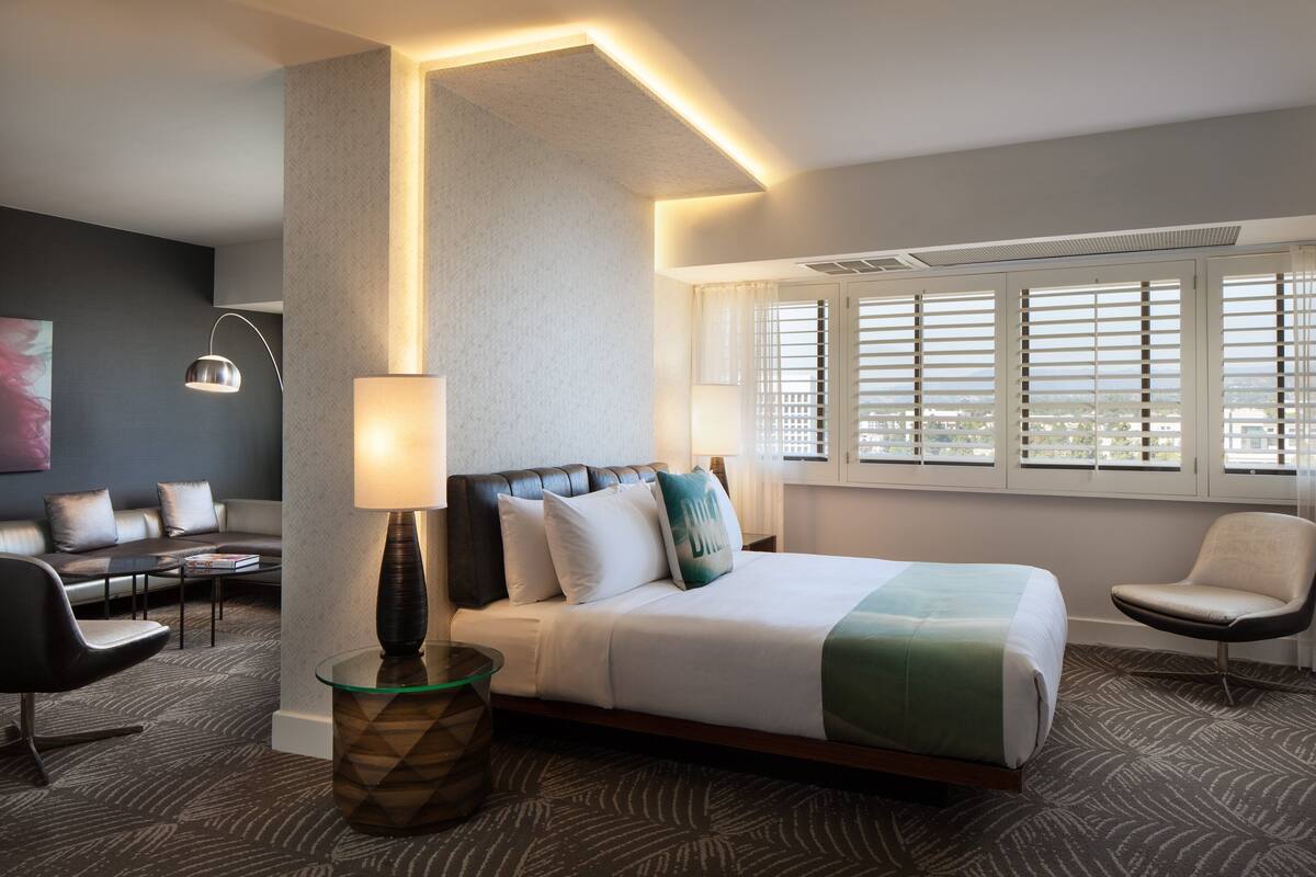 Une chambre d’hôtel équipée d’un lit, d’un bureau, d’une chaise, d’une lampe et d’une fenêtre avec des stores.