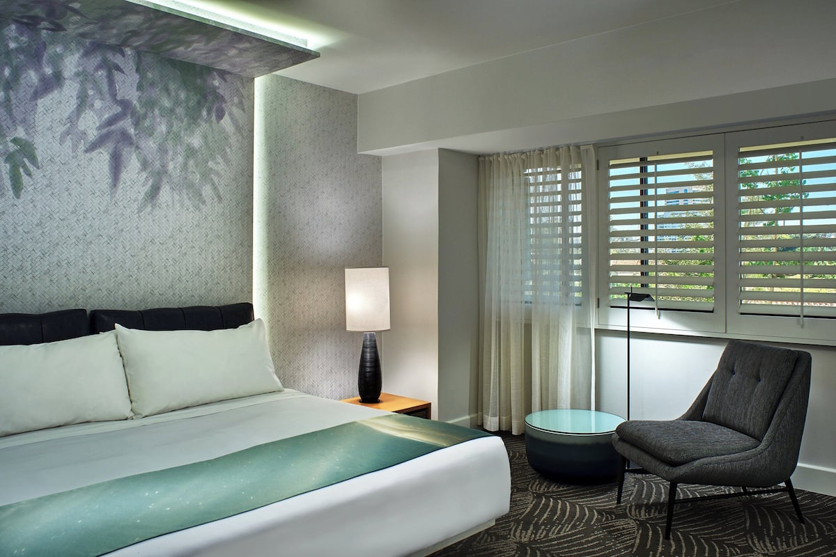 Une chambre d’hôtel avec un lit, une chaise, une lampe et une vue sur la verdure par la fenêtre.