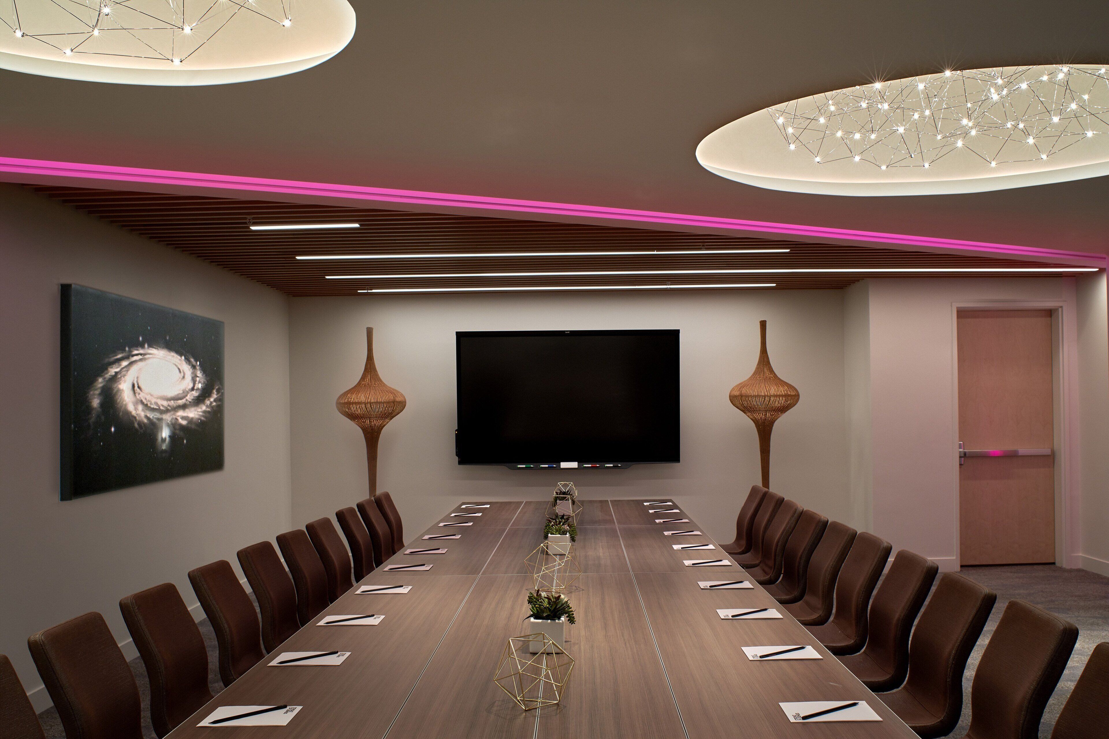 A conference room with a long table, brown chairs, a flat-screen TV, and modern lighting fixtures.