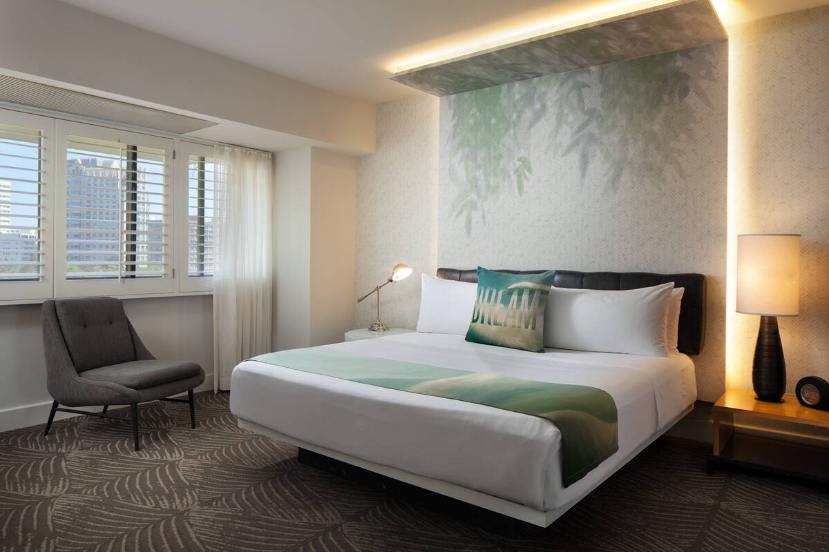 Une chambre d’hôtel avec un grand lit, une chaise, une table de chevet avec une lampe et une fenêtre avec des stores.