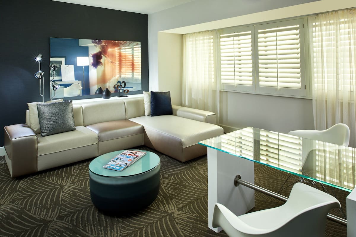 Une chambre d’hôtel moderne avec un canapé d’angle, une table basse en verre, un fauteuil blanc et une grande fenêtre avec des stores.