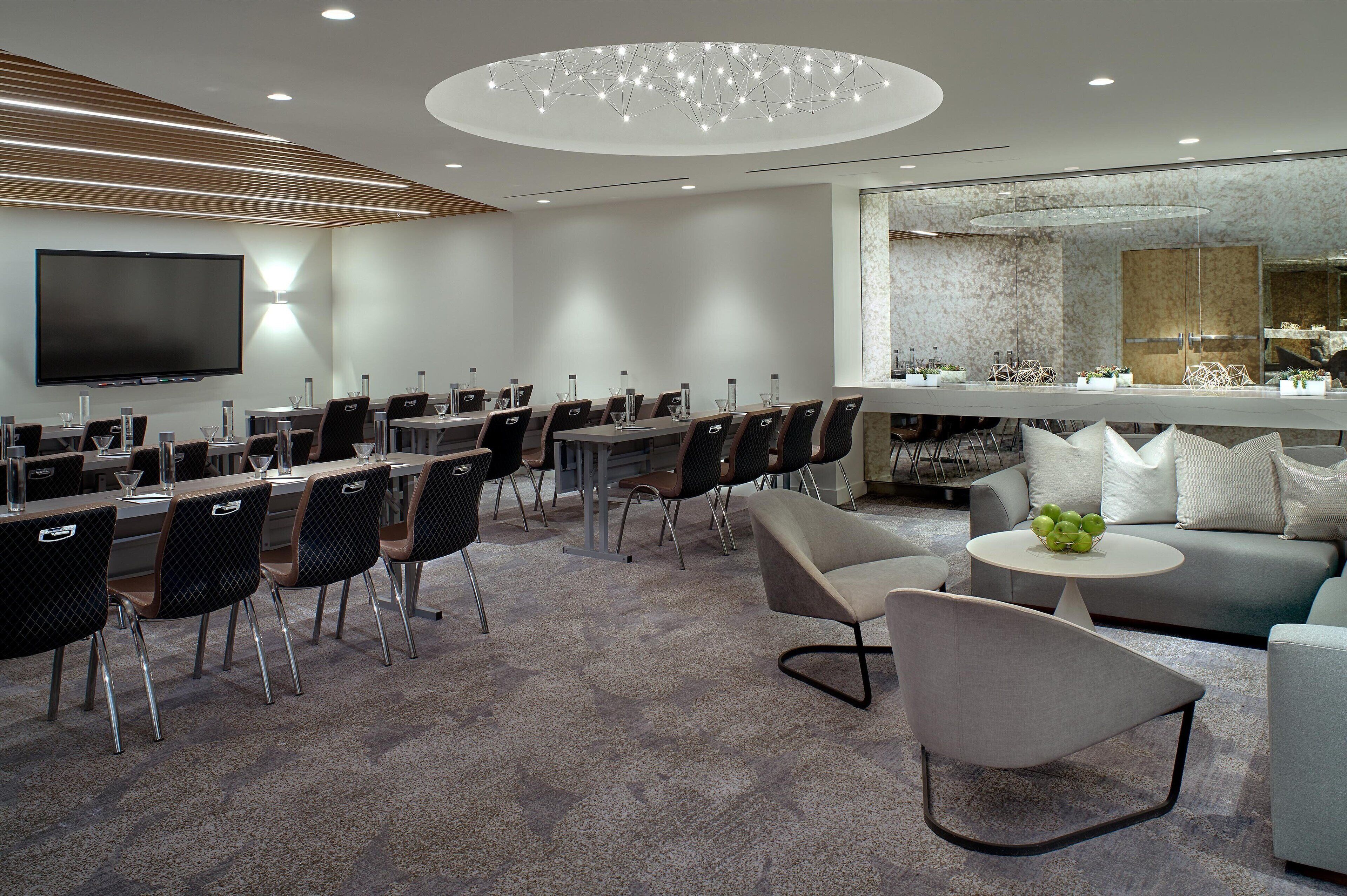 A modern hotel lobby with a long dining table, multiple chairs, a flat-screen TV, and a seating area with a sofa and coffee table.