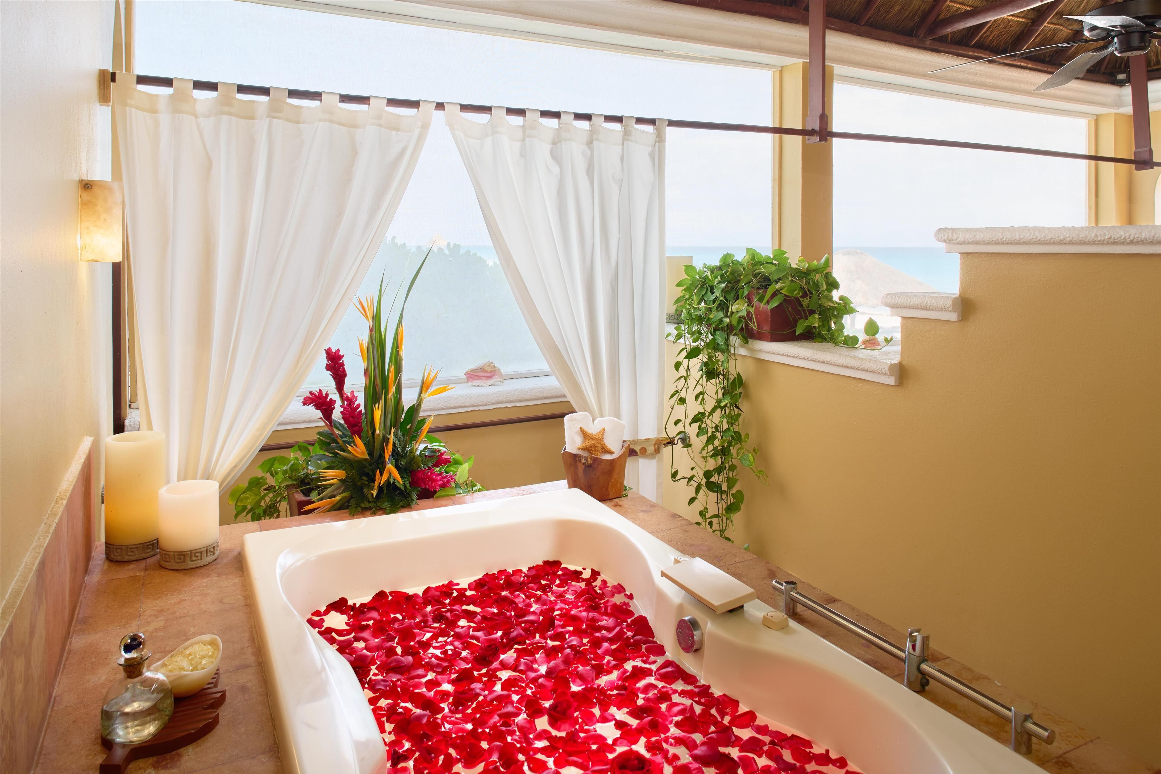 A bathroom with a bathtub filled with rose petals, a window with curtains, and a view of the sea.
