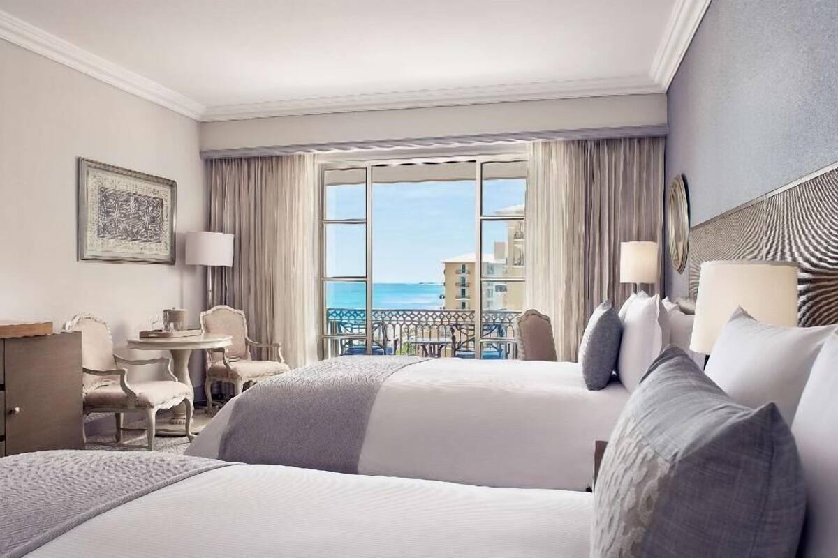 Une chambre d’hôtel avec deux lits, un bureau, une chaise, une petite table et un balcon donnant sur la mer.