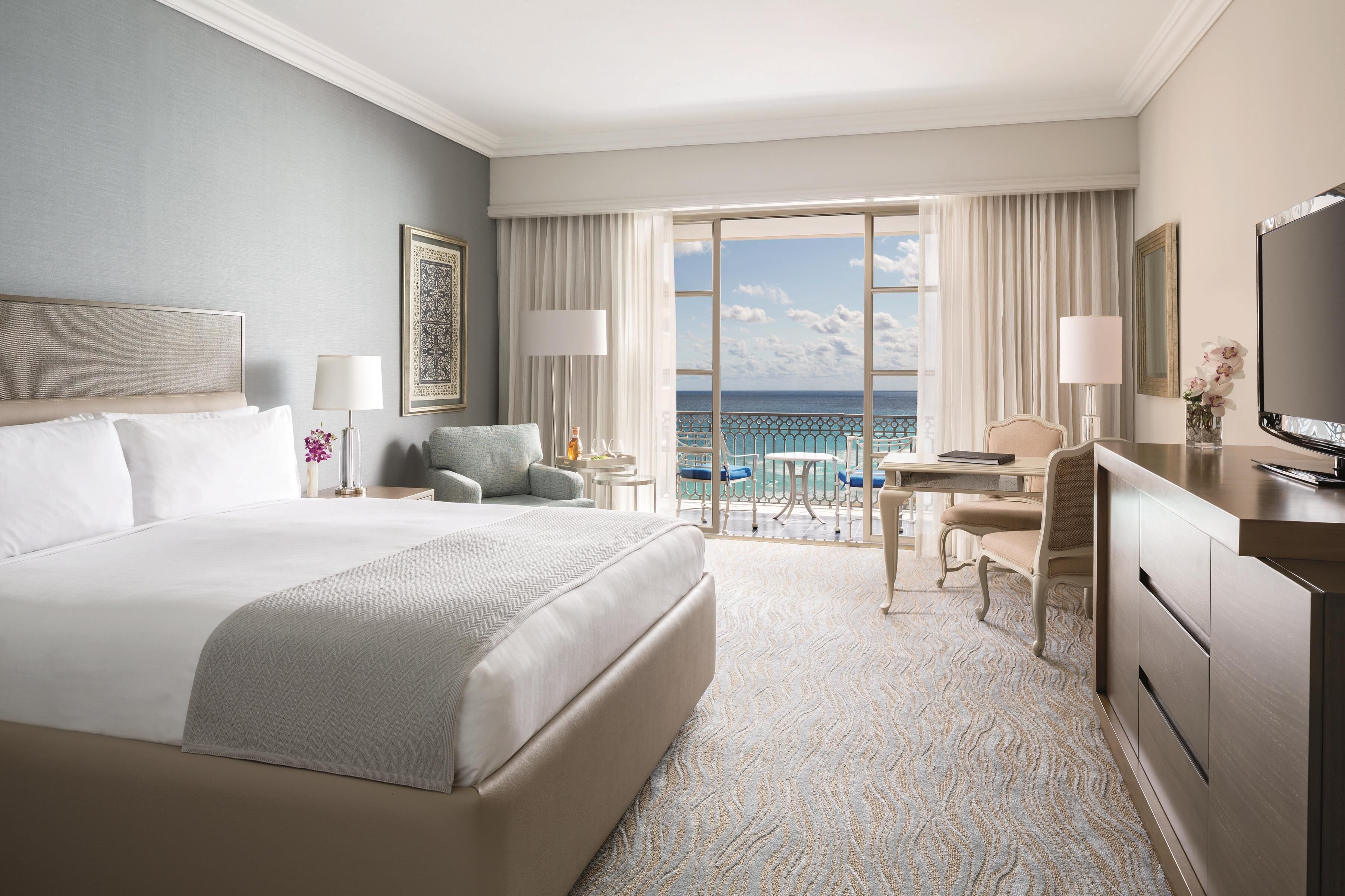 A hotel room with a large bed, a desk, a chair, a TV, and a balcony with a view of the ocean.